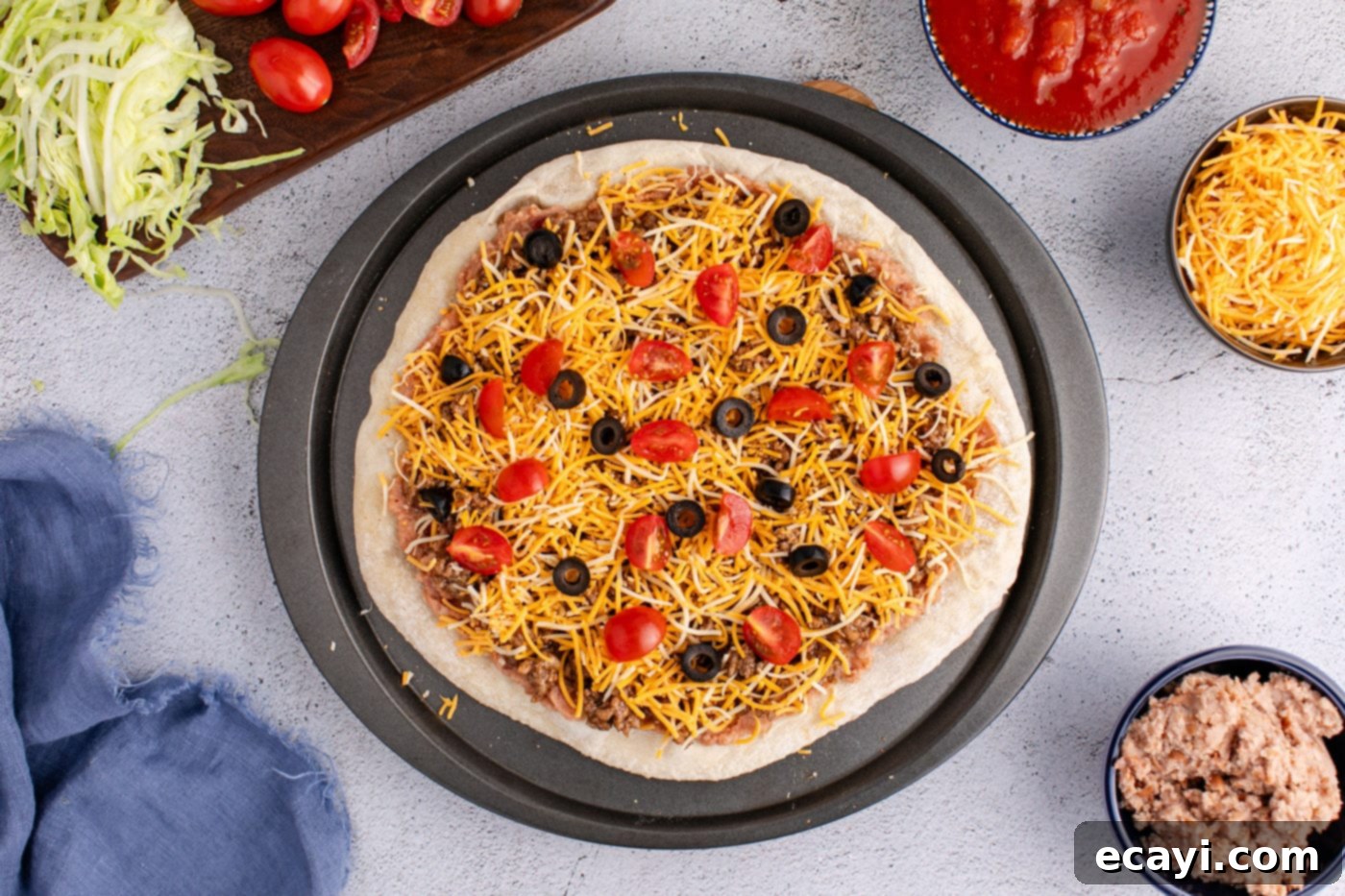 Fiesta Taco Pizza 8 Taco pizza partially assembled with shredded cheese, taco meat, tomatoes, and olives on top of the refried bean and pizza crust base, ready for baking.