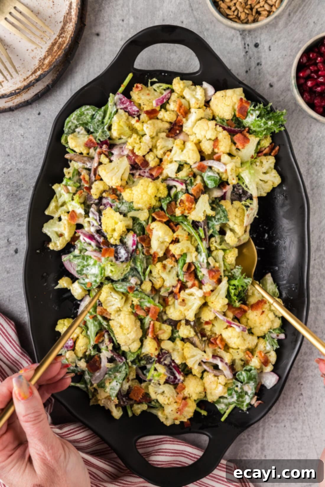 Zesty Cauliflower Medley 10 Hands picking up a generous portion of Creamy Roasted Cauliflower Salad using serving spoons, showcasing its appealing texture.
