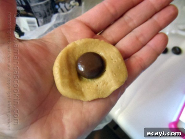 Chocolate-Stuffed Peanut Butter Perfection 5 A hand gently flattens a portion of peanut butter cookie dough in its palm, preparing to place a shiny Hershey's Drop chocolate candy in the center. The dough appears soft and pliable, ready for its sweet filling.