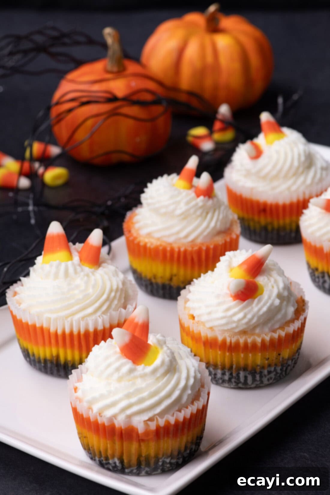 Festive Candy Corn Mini Cheesecakes 2 A trio of colorful Mini Candy Corn Cheesecakes presented on a white platter, with decorative pumpkins softly blurred in the background, ready for a Halloween celebration.