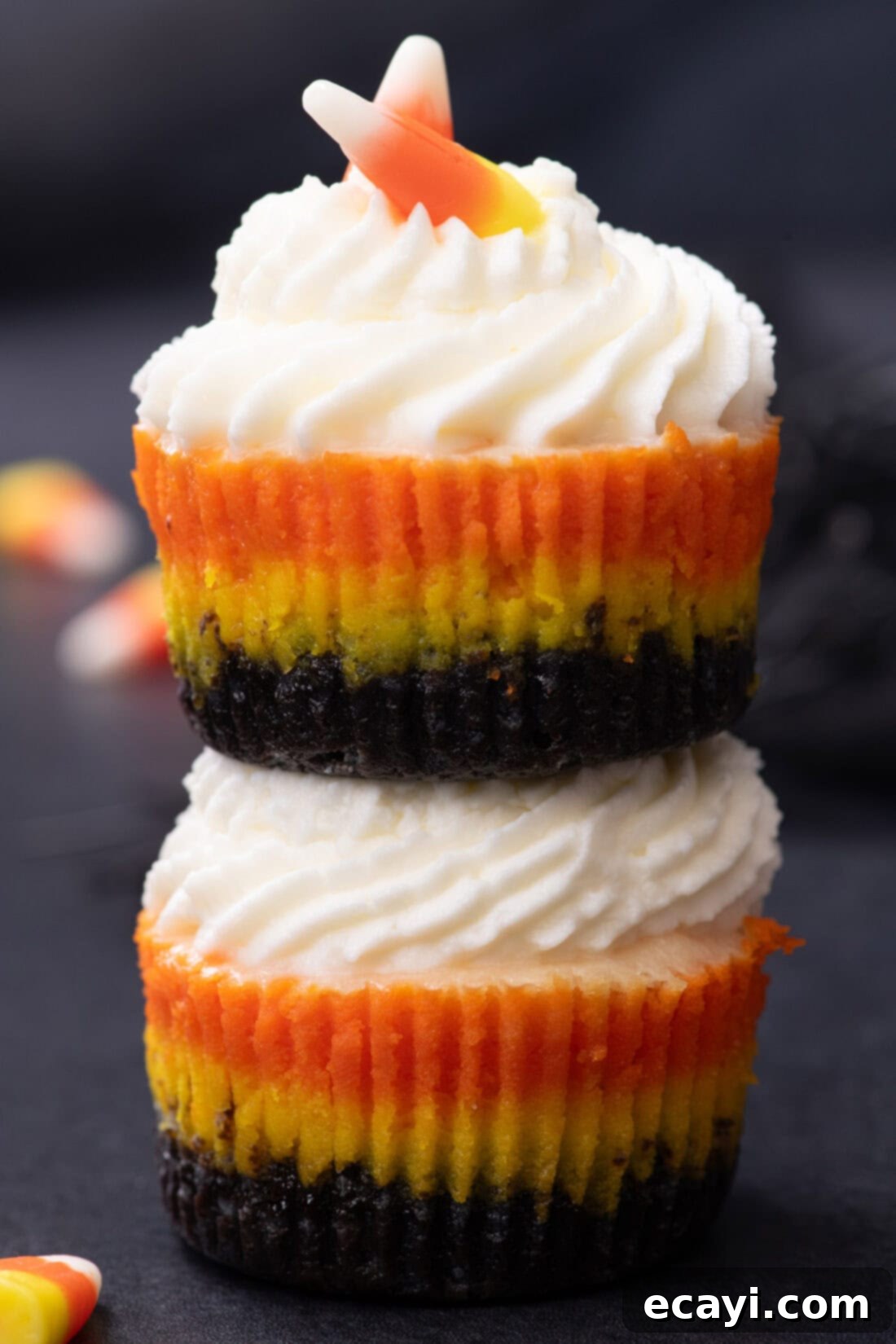 Festive Candy Corn Mini Cheesecakes 3 Two delightful Mini Candy Corn Cheesecakes, each adorned with a swirl of whipped cream and a piece of candy corn, are stacked on top of each other, showcasing their vibrant layers.