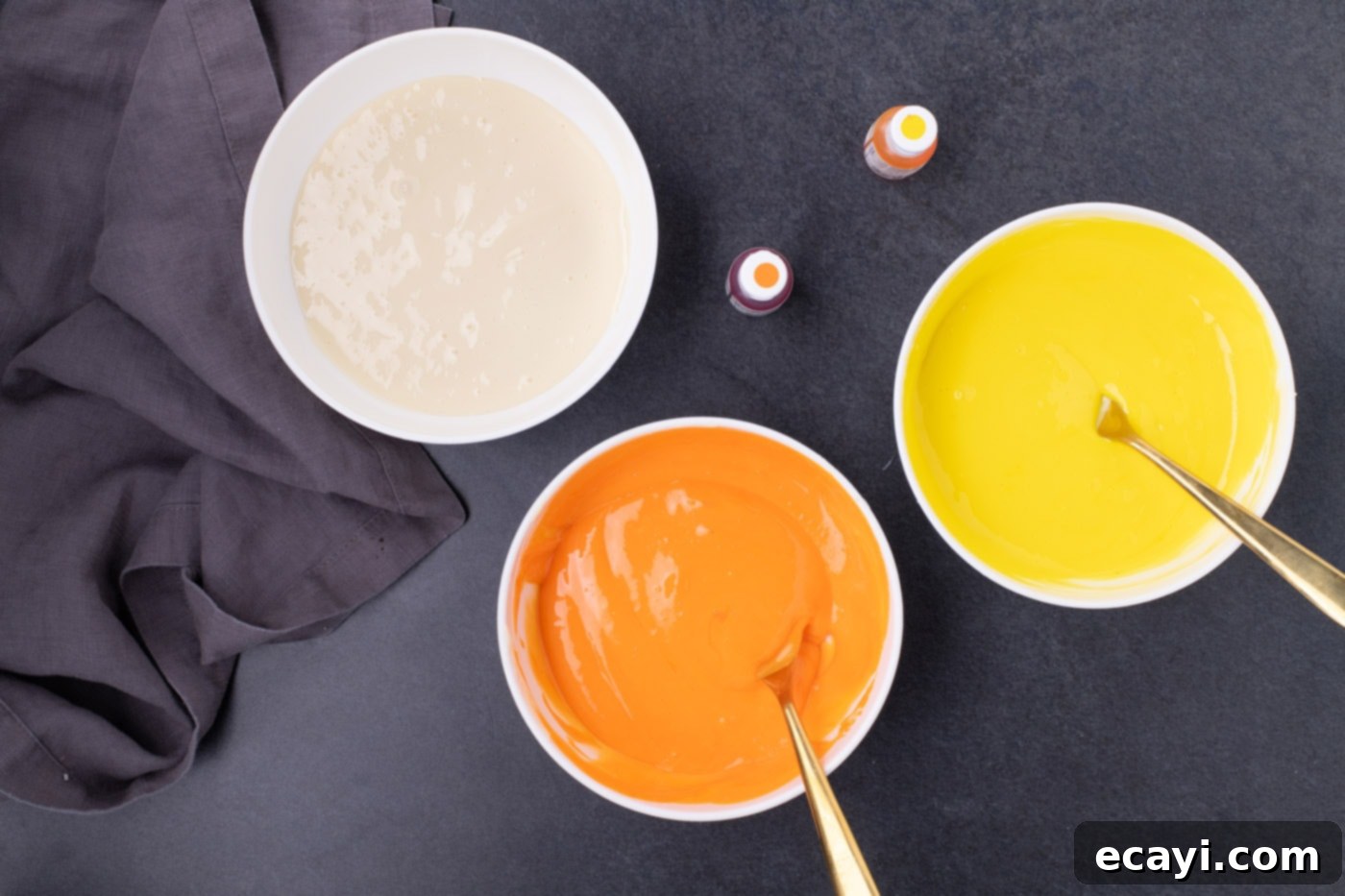 Festive Candy Corn Mini Cheesecakes 9 Three bowls of cheesecake filling, one yellow, one orange, and one cream-colored, showcasing the vibrant shades achieved with gel food coloring.