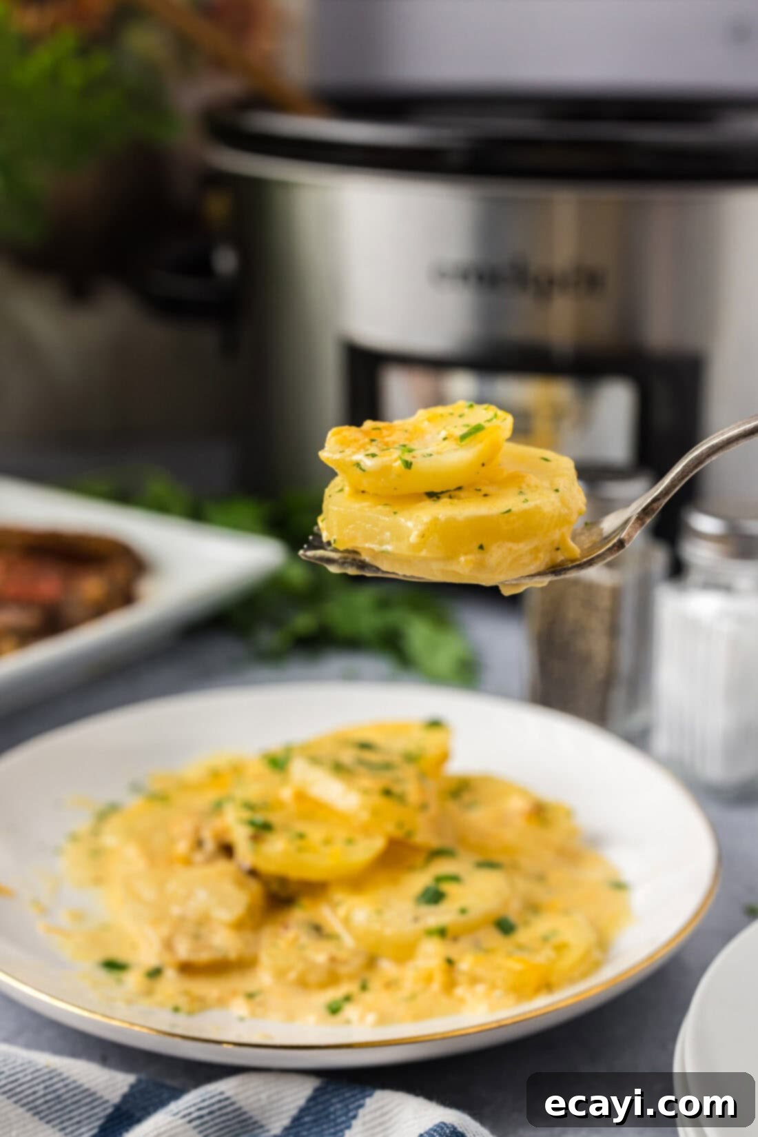 Set It and Forget It Scalloped Potatoes 2 Two Crockpot Scalloped Potatoes on a fork held above a plate of Crockpot Scalloped Potatoes