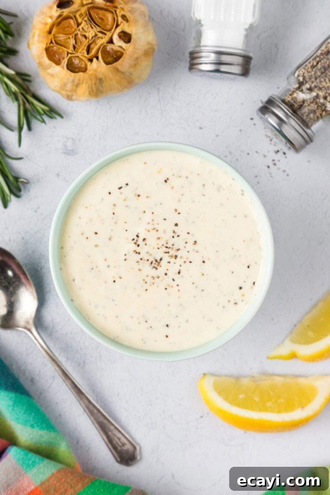 Creamy Garlic Aioli 7 A beautifully arranged bowl of creamy Roasted Garlic Aioli, garnished with fresh herbs and surrounded by lemons, garlic cloves, salt, and pepper.