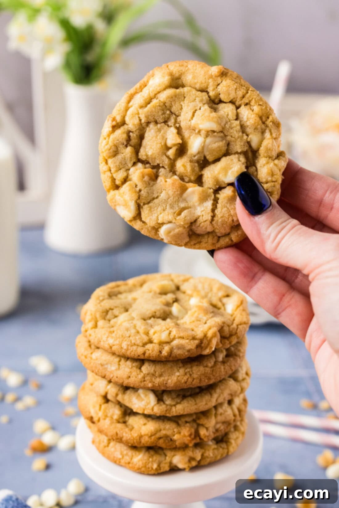 Decadent White Chocolate Macadamia Delights 2 A hand holding up a freshly baked, perfectly golden-brown White Chocolate Macadamia Nut Cookie, with a stack of more delicious cookies blurred in the background, showcasing the irresistible texture and generous fillings.