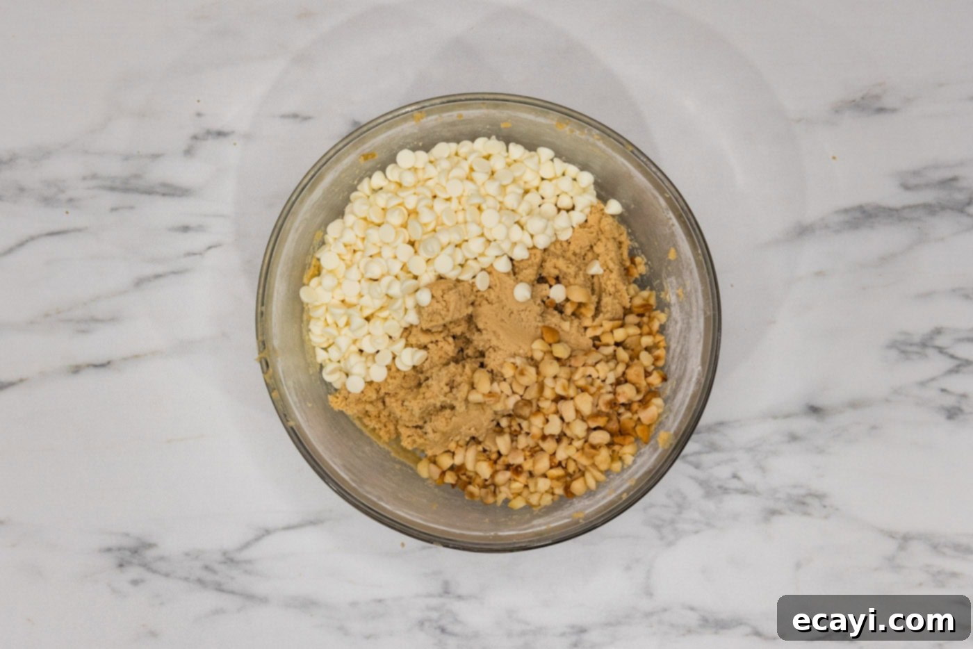 Decadent White Chocolate Macadamia Delights 8 White chocolate chips and macadamia nuts freshly added to a bowl of cookie dough, ready to be folded in with a spatula for even distribution.
