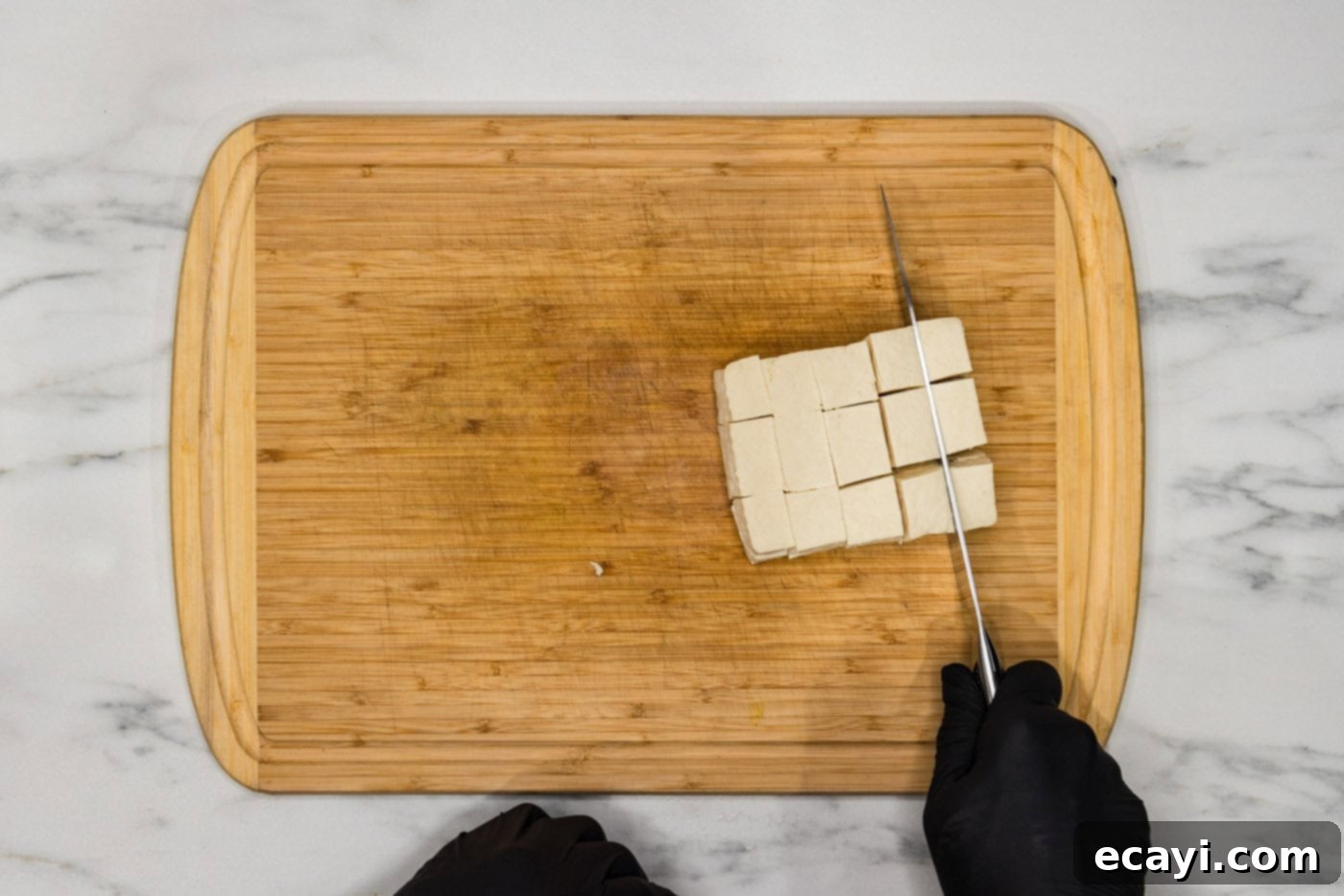 Crispy Golden Tofu 6 A sharp chef's knife precisely slicing a pressed block of firm tofu into uniform 2-inch cubes on a clean wooden cutting board, ready for seasoning.
