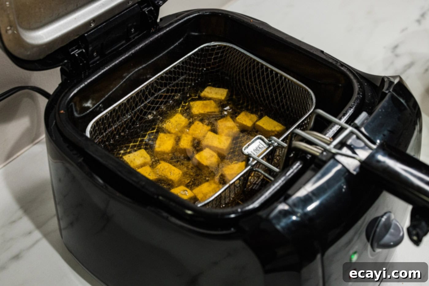 Crispy Golden Tofu 9 Tofu cubes actively frying in a deep fryer basket, fully submerged in hot oil, steadily transforming into a beautiful golden brown color with a developing crispy crust.