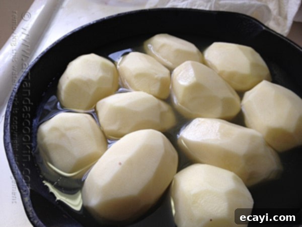 Crispy Cast Iron Roast Potatoes 6 Peeled potatoes in a cast iron skillet with broth and thin slices of cold butter on top, poised for roasting.