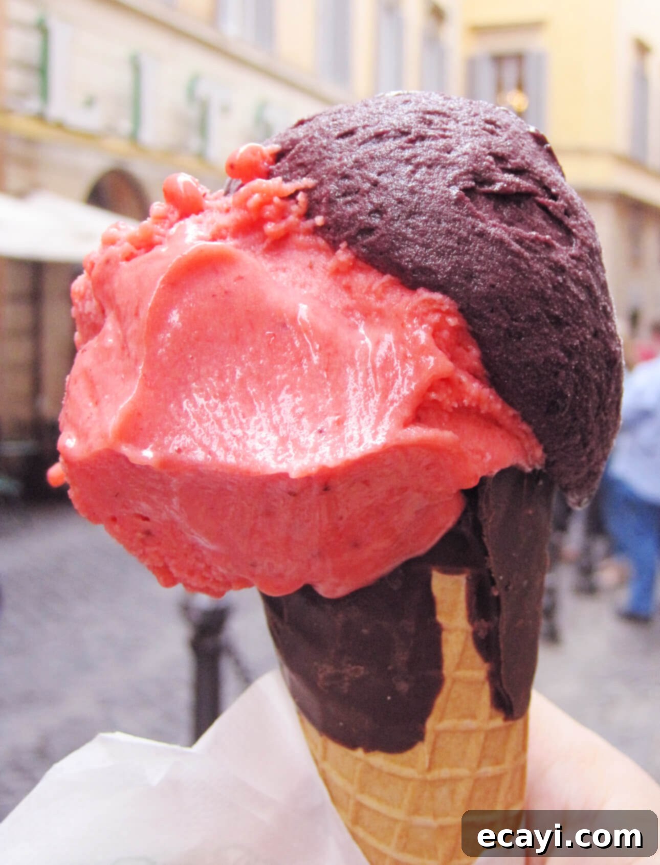 Velvet Blueberry Gelato 4 A hand holds an ice cream cone featuring two scoops—rich blueberry and vibrant pink strawberry, with the cone partially dipped in chocolate. A hint of blueberry gelato swirls around. The background reveals a blurred street scene, bustling with buildings and people. // FoodNouveau.com