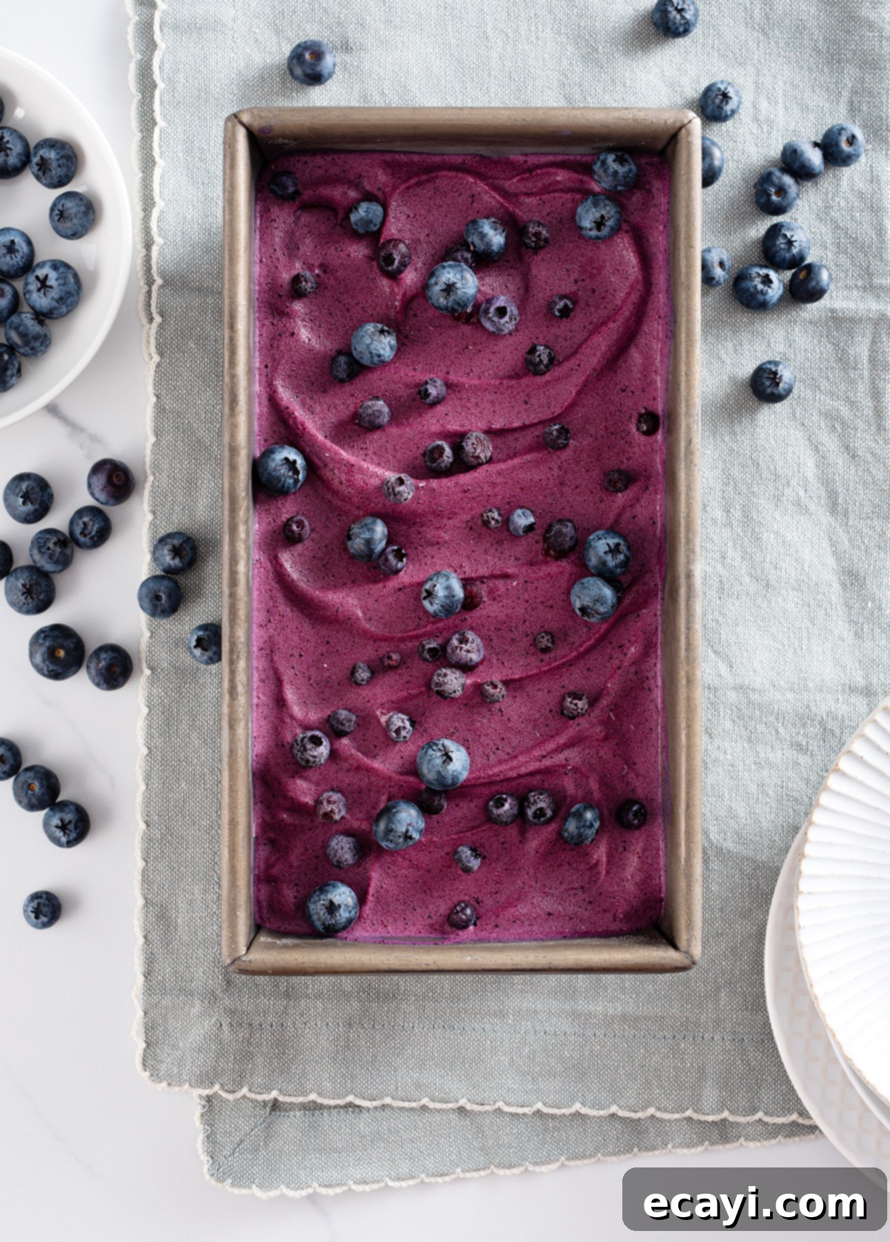 Velvet Blueberry Gelato 5 A rectangular pan filled with swirled blueberry gelato topped with fresh blueberries. Surrounding the pan are scattered blueberries, a small bowl of blueberries, and a white plate on a light gray tablecloth. // FoodNouveau.com