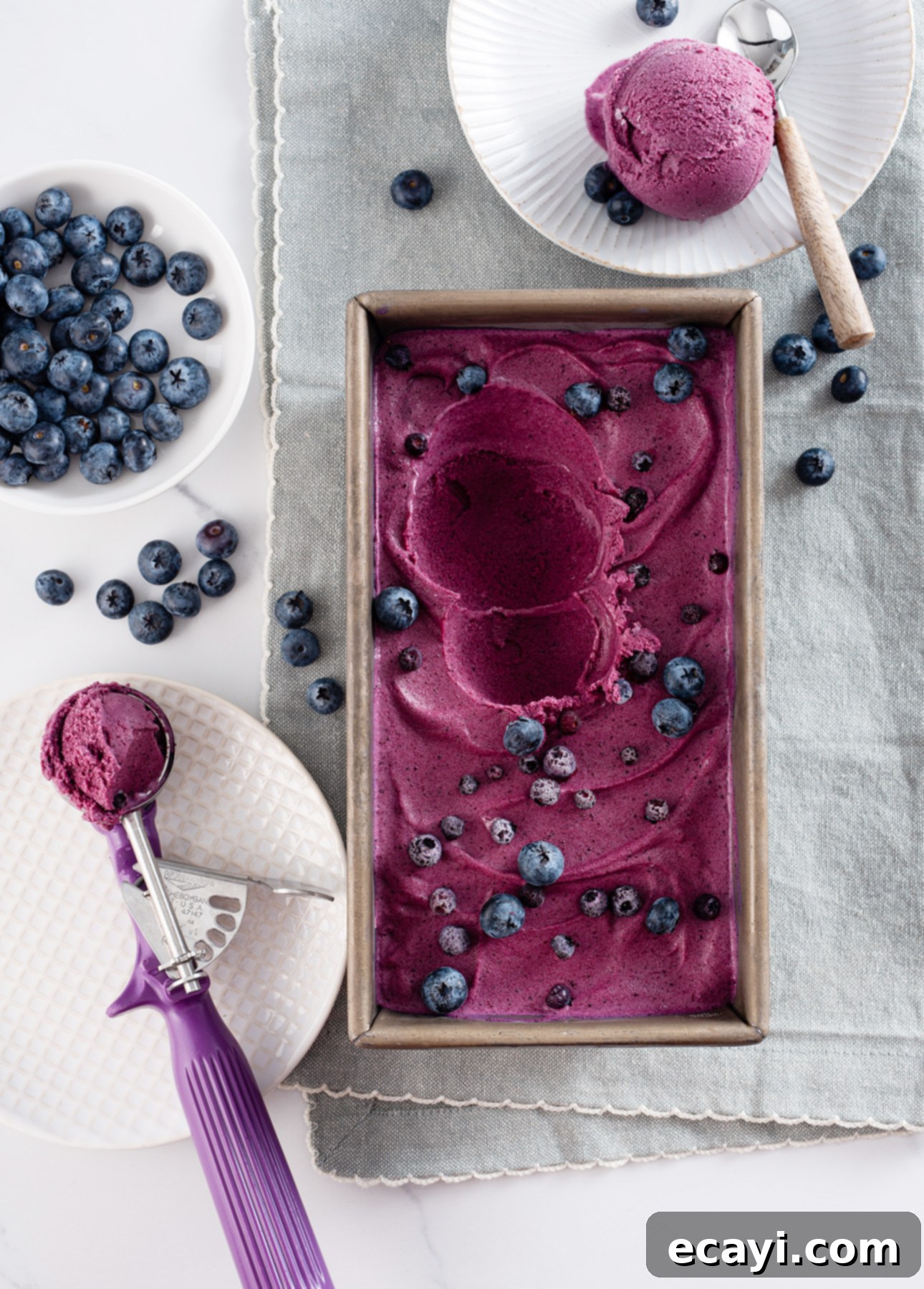 Velvet Blueberry Gelato 7 A rectangular dish filled with vibrant purple blueberry gelato is topped with fresh blueberries. Next to it are plates each boasting a scoop of this delightful gelato and more blueberries. A purple ice cream scoop rests beside the dish on a textured plate. // FoodNouveau.com