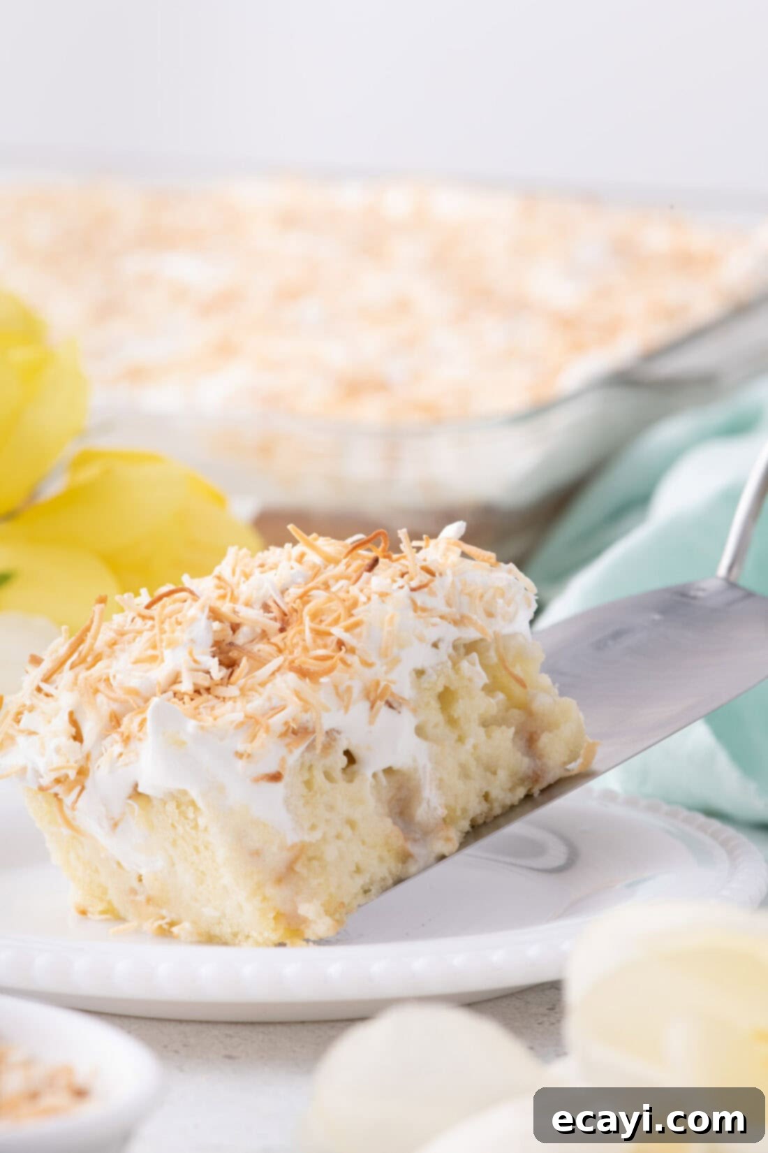 Tropical Coconut Infusion Cake 2 Slice of Coconut Poke Cake being served onto a plate, showcasing its layers and toasted coconut topping.