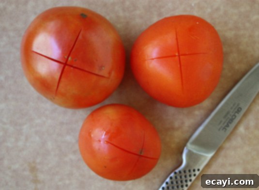 How to peel fresh tomatoes: Slice a shallow "X" on the bottom of each tomato // FoodNouveau.com Close-up of a red tomato with a small "X" scored into its skin at the bottom, indicating preparation for peeling. Essential for homemade fresh tomato sauce.