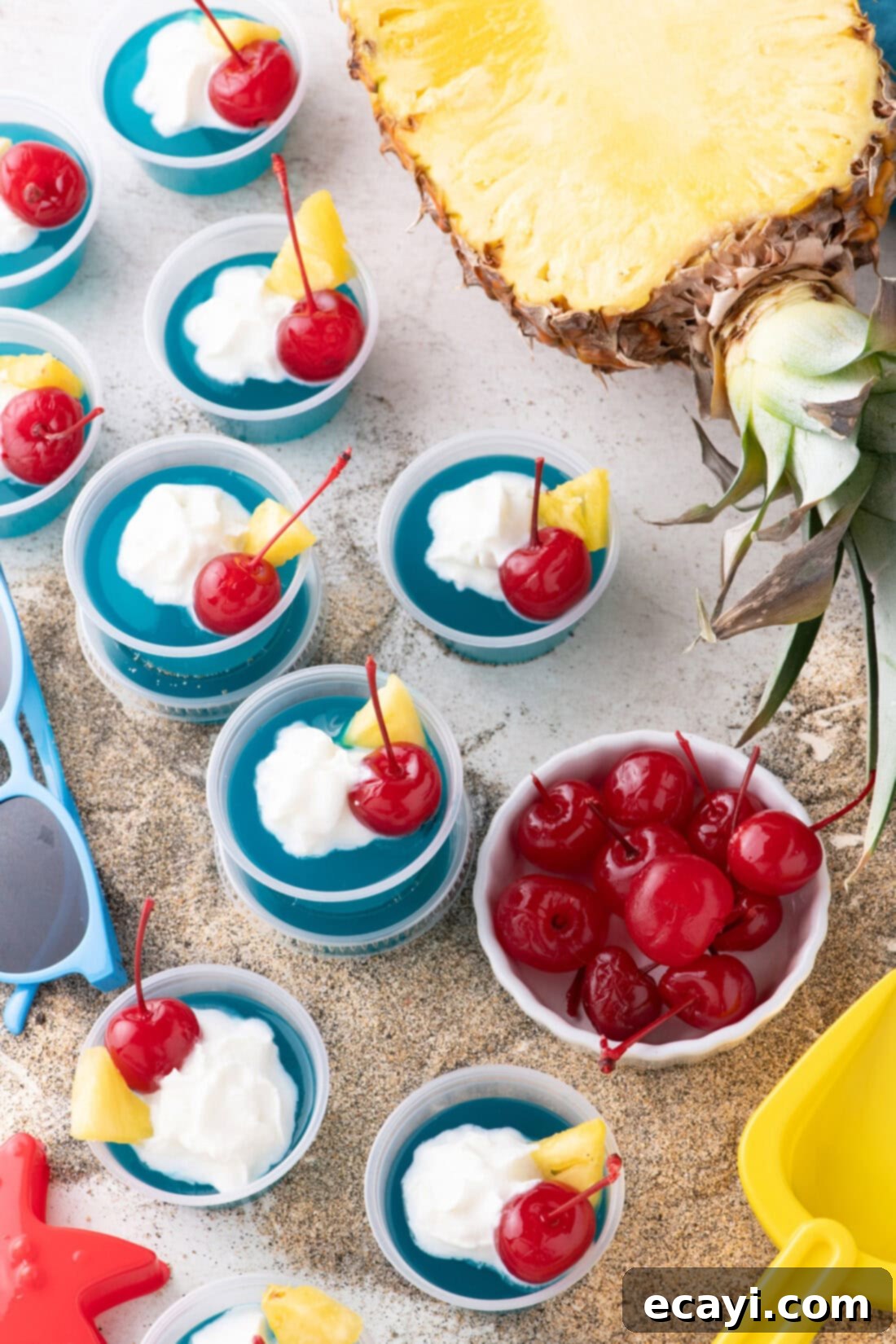 Blue Lagoon Jello Shooters 2 Vibrant Blue Hawaiian Jello Shots resting on sand with a fresh pineapple and maraschino cherries in the background, evoking a tropical beach scene.