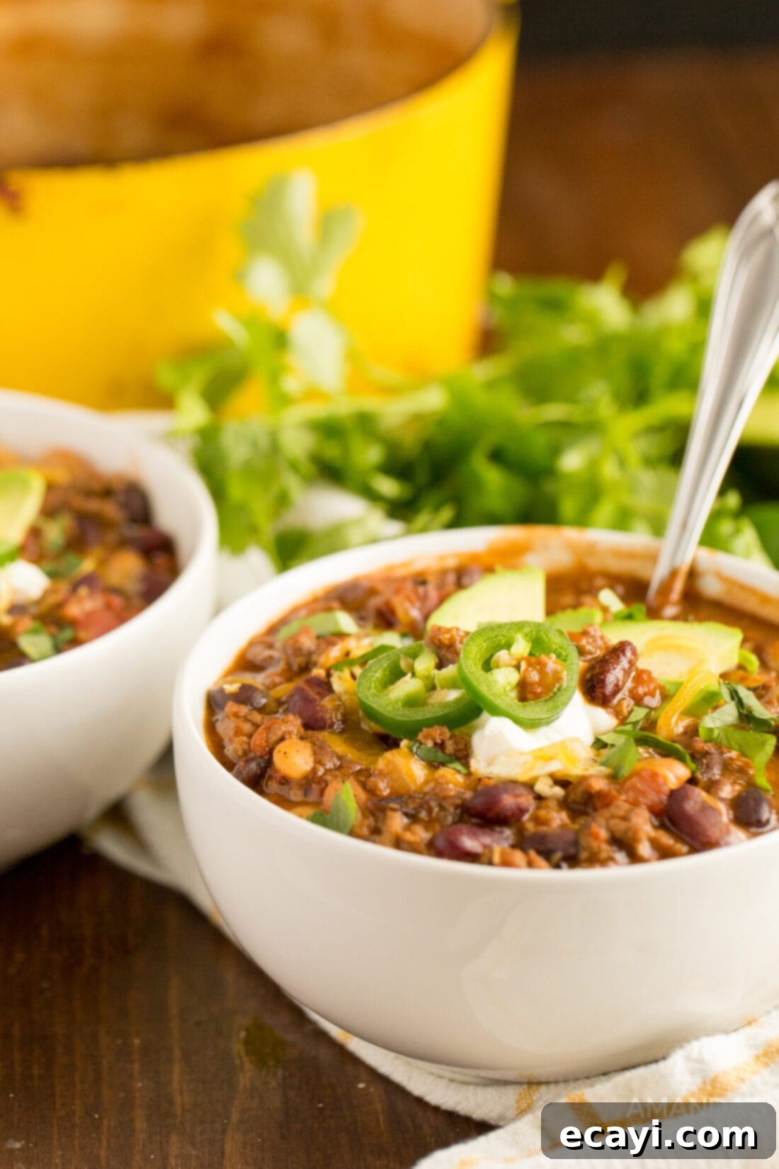This 3 Bean Chili is made with a homemade chili seasoning, fresh ground beef, fire roasted tomatoes, and three types of canned beans. Great for football!  Two steaming bowls of hearty 3 Bean Chili garnished with fresh herbs and shredded cheese, inviting you to dig in.