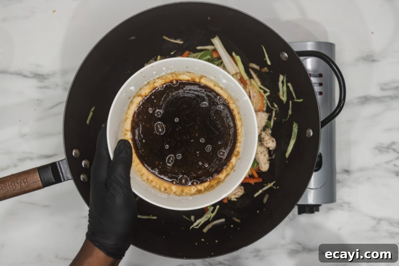 Classic Chicken Lo Mein 9 gloved hand pouring lo mein sauce from a bowl into a wok containing chicken and vegetables