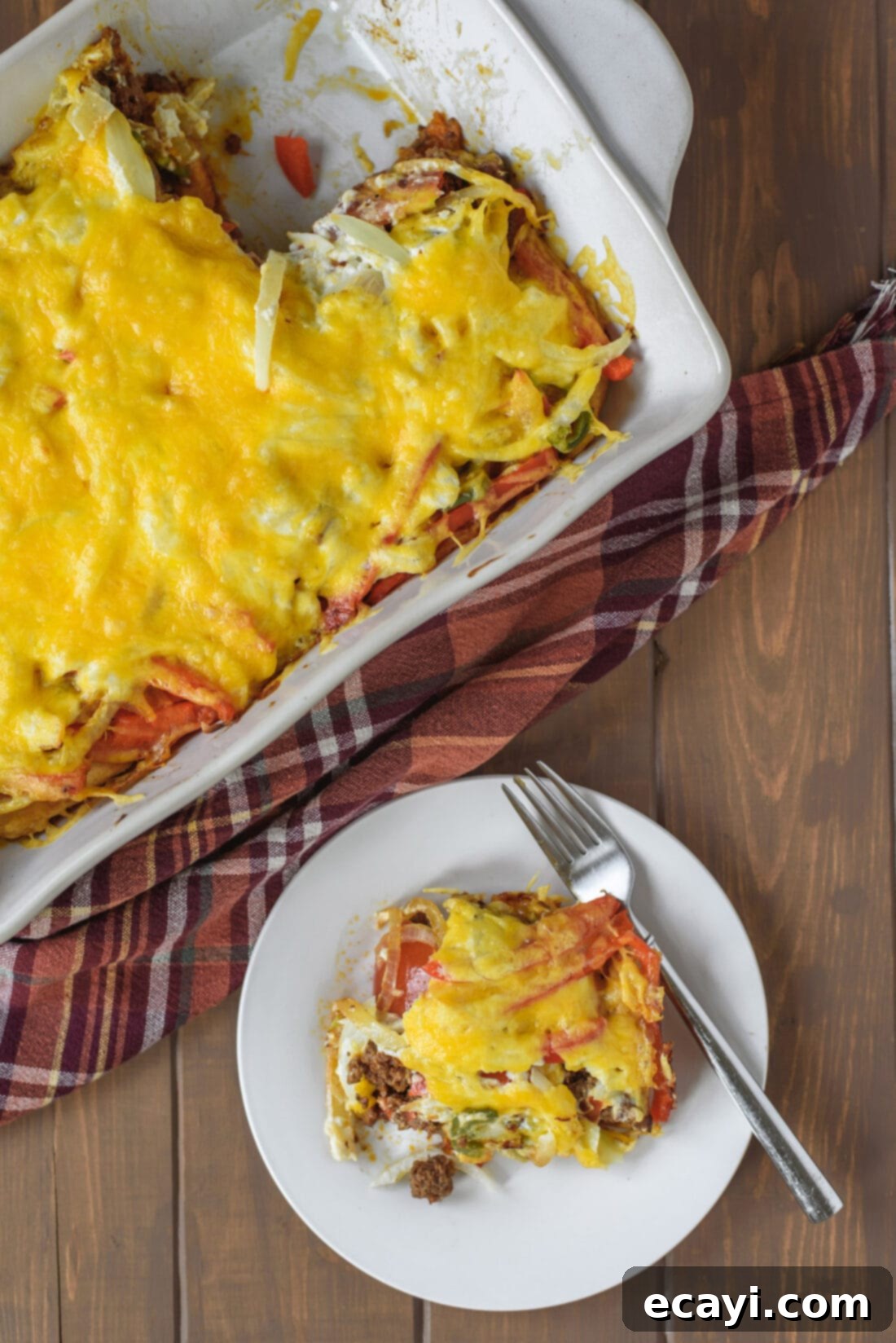 John Wayne Casserole: A delicious beef and biscuit casserole with cheese, fresh vegetables, and a hint of jalapeño. Perfect for dinner! A slice of John Wayne Casserole on a plate next to the baking pan, showing off its delicious layers of beef, cheese, and vegetables on a biscuit crust.