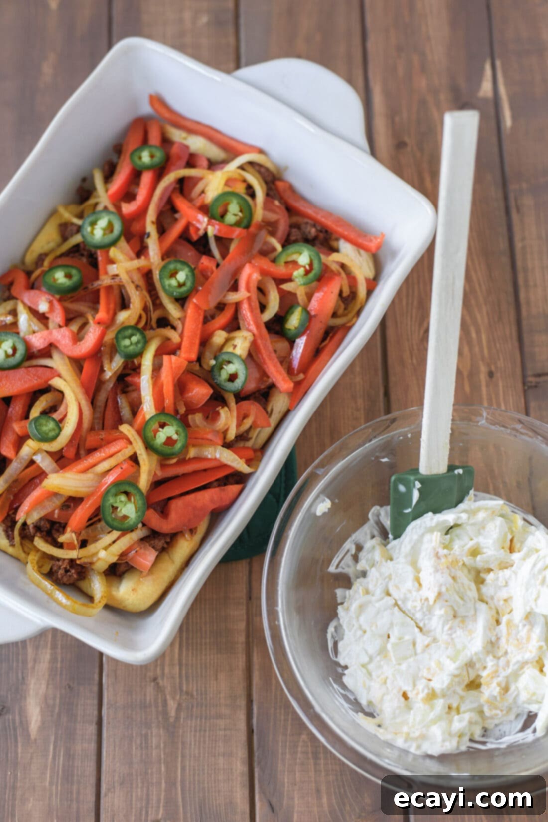 Layering the delicious John Wayne Casserole with the creamy cheese mixture ready to go! A layered John Wayne Casserole in a baking dish, before baking, next to a bowl of the creamy cheese mixture.