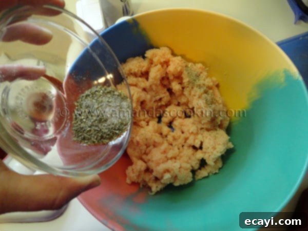 homemade-chicken-nuggets-step-4 A hand gently sprinkling a mix of salt, pepper, onion powder, oregano, and parsley over the processed raw chicken in a white mixing bowl.