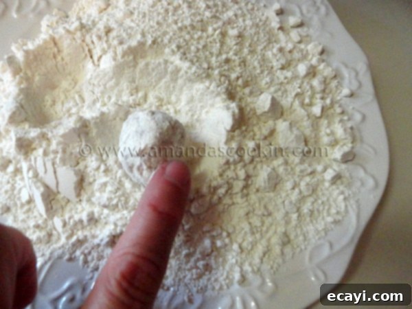 homemade-chicken-nuggets-step-9 A raw chicken ball being gently rolled and coated in the flour mixture on a white plate, preparing it for the next step.