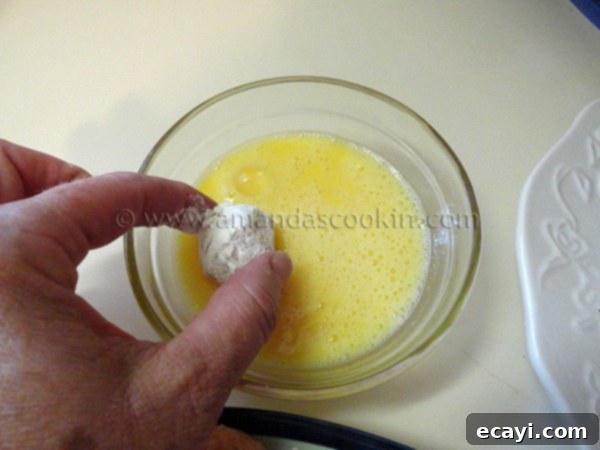 homemade-chicken-nuggets-step-10 A close-up of a floured chicken ball being dipped into and coated with whisked egg in a clear glass bowl, showcasing the wet coating step.