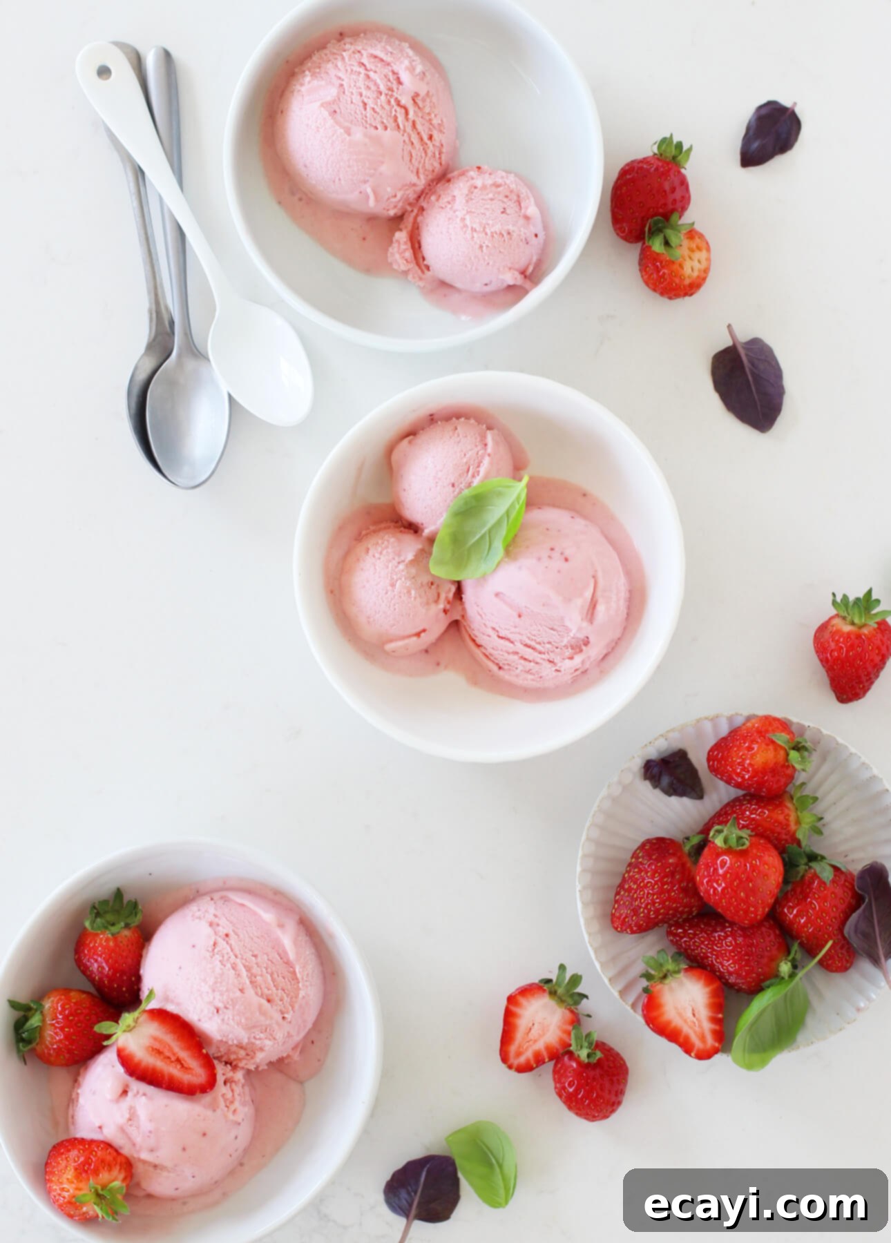 Basil-Kissed Strawberry White Chocolate Gelato 2 Lush pink strawberry gelato bowls, garnished with fresh strawberries and vibrant basil leaves, resting on a pristine white surface alongside a small dish of berries and three spoons. This white chocolate, strawberry, and basil gelato is a perfect summer dessert from FoodNouveau.com.