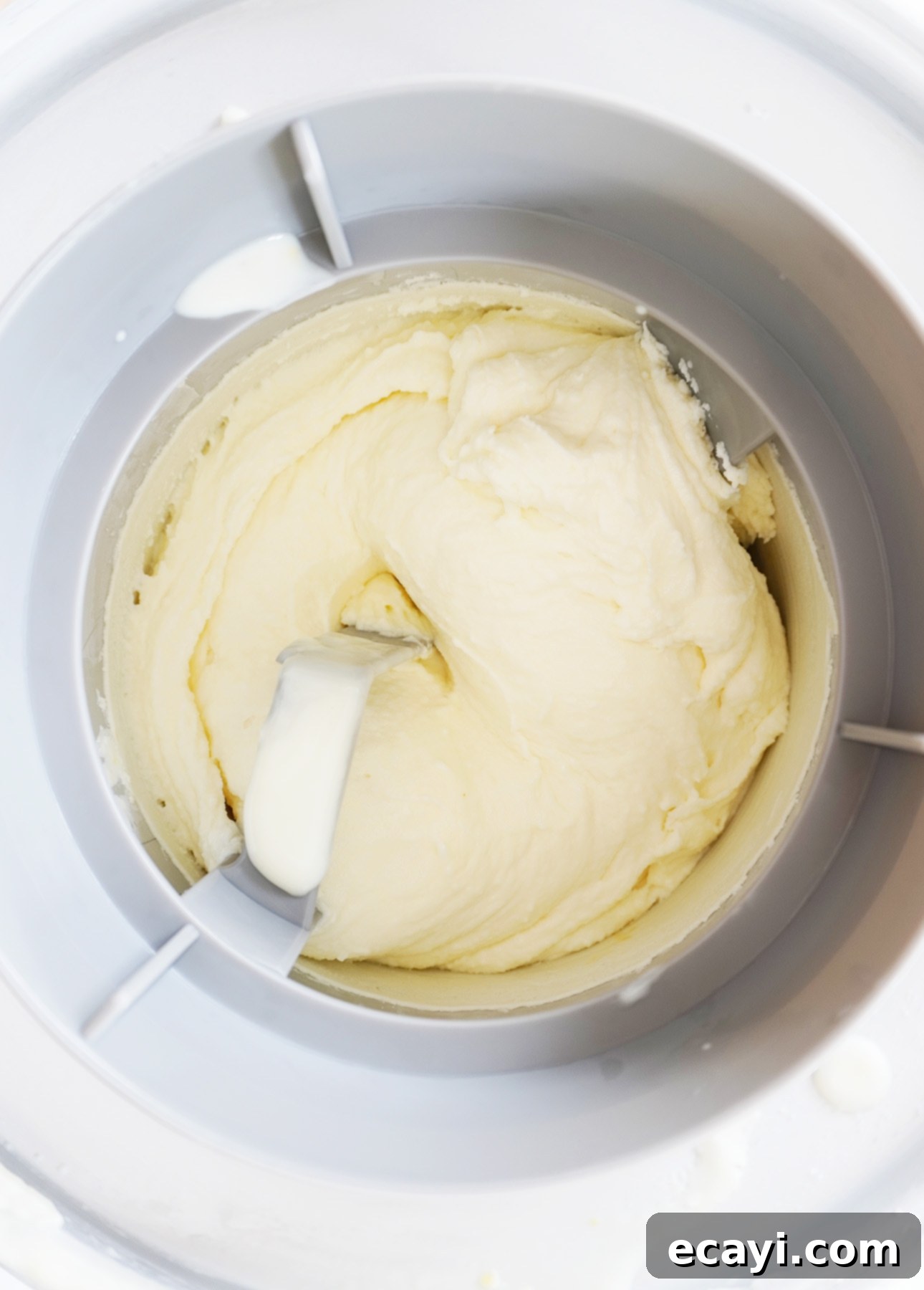 Crafting Creamy Vegan Gelato at Home 4 Close-up of creamy gelato being churned in a white ice cream maker. A metal paddle is visible, artfully mixing the smooth texture within the machine, creating a luscious base for homemade vegan gelato. // FoodNouveau.com