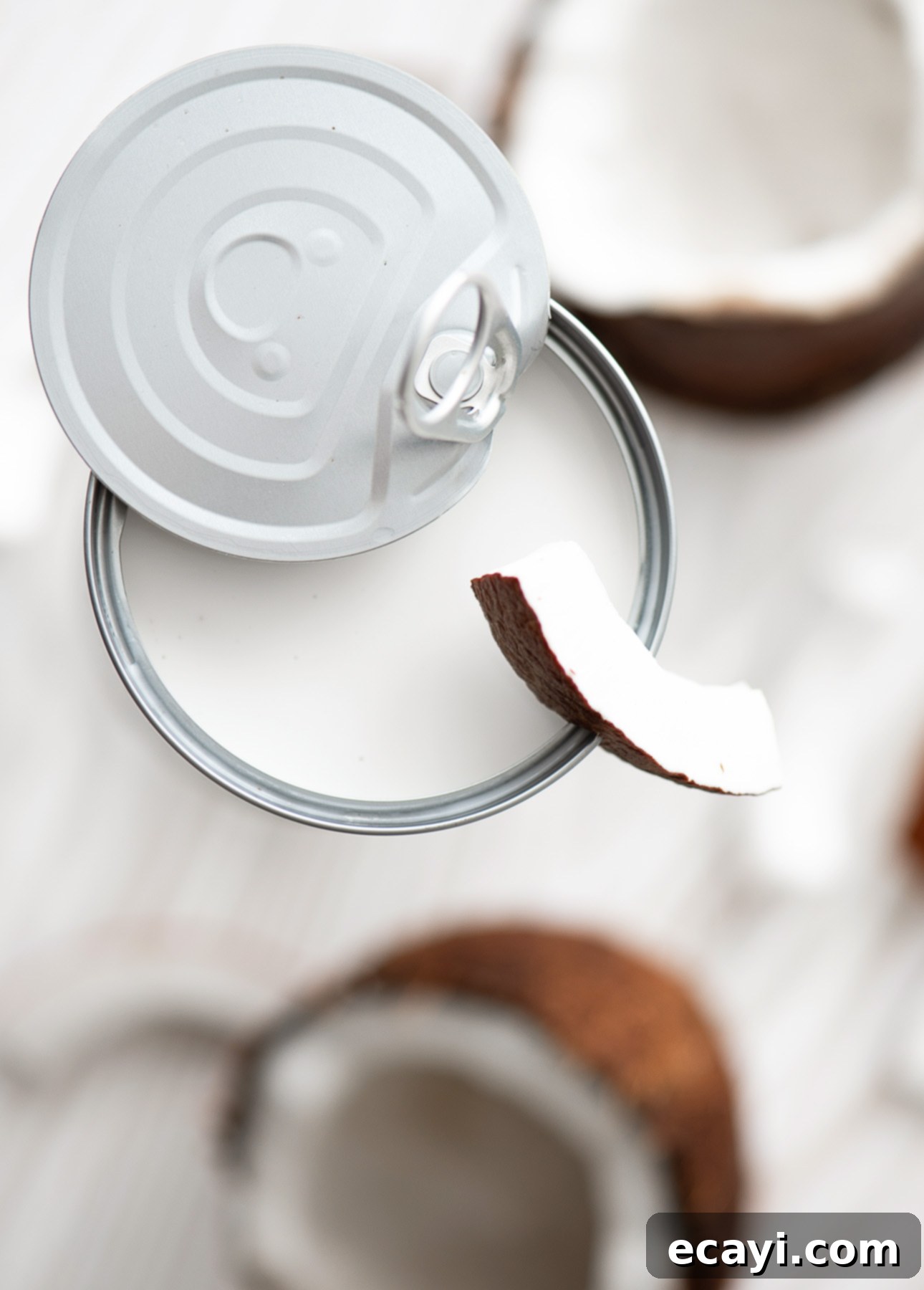 Crafting Creamy Vegan Gelato at Home 6 A top view of an opened can of coconut milk with the lid pulled back, a slice of fresh coconut perched on the edge. Two blurred coconut halves linger in the background on a white surface, perfect for homemade plant-based gelato. // FoodNouveau.com
