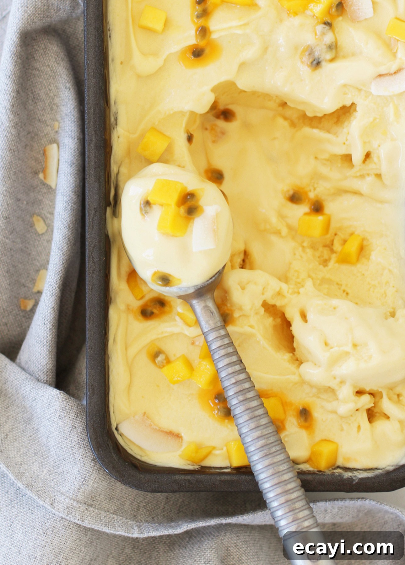 Crafting Creamy Vegan Gelato at Home 8 A close-up of creamy yellow mango vegan gelato adorned with chunks of mango and passion fruit seeds, elegantly served in a metal container, showing its rich, dairy-free texture. // FoodNouveau.com