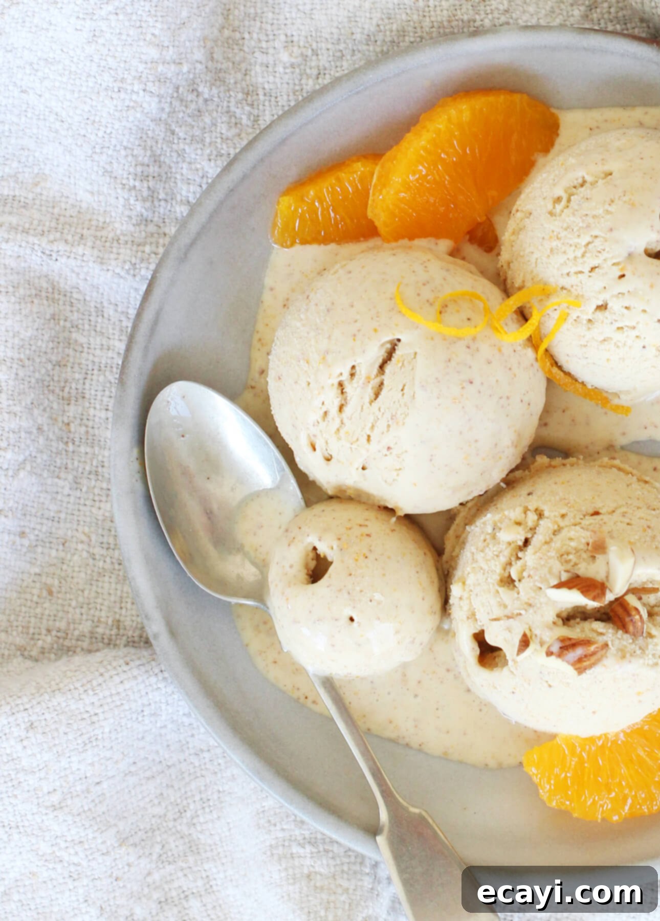Sun-Kissed Orange Almond Gelato 3 A plate of orange almond gelato with three scoops, garnished with orange slices and a twist of peel. A spoon rests beside the scoops on a light-colored cloth, creating a cozy setting. // FoodNouveau.com