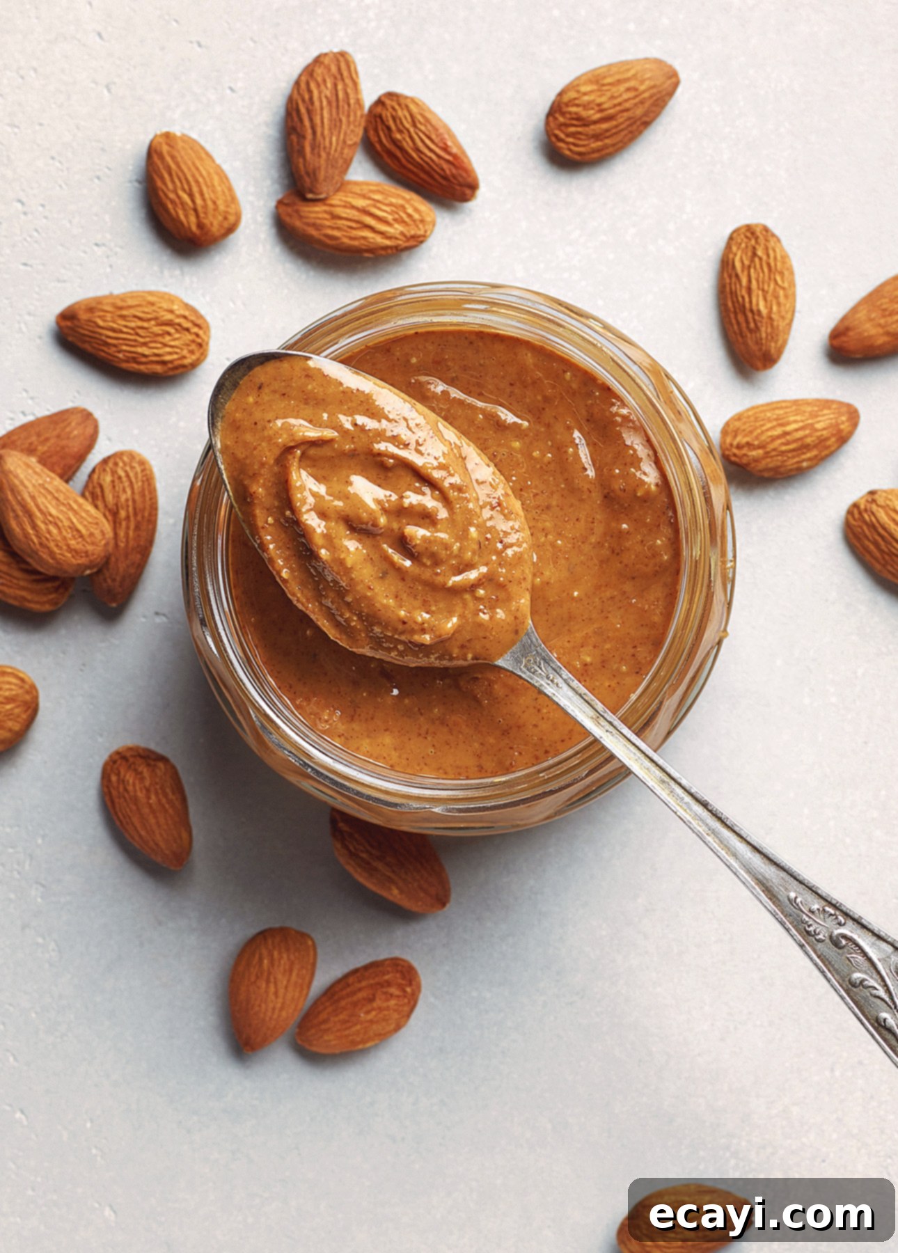 Sun-Kissed Orange Almond Gelato 4 A spoonful of almond butter is held above an open jar filled with the spread. Raw almonds are scattered around the jar on a white surface. // FoodNouveau.com