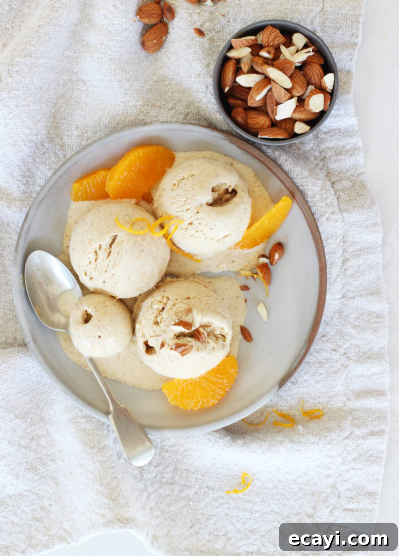 Sun-Kissed Orange Almond Gelato 5 A bowl of orange almond gelato is garnished with almond pieces and fresh orange slices, elegantly placed on a textured napkin. Nearby, a small bowl holds mixed almonds, while a spoon rests invitingly on the plate. // FoodNouveau.com