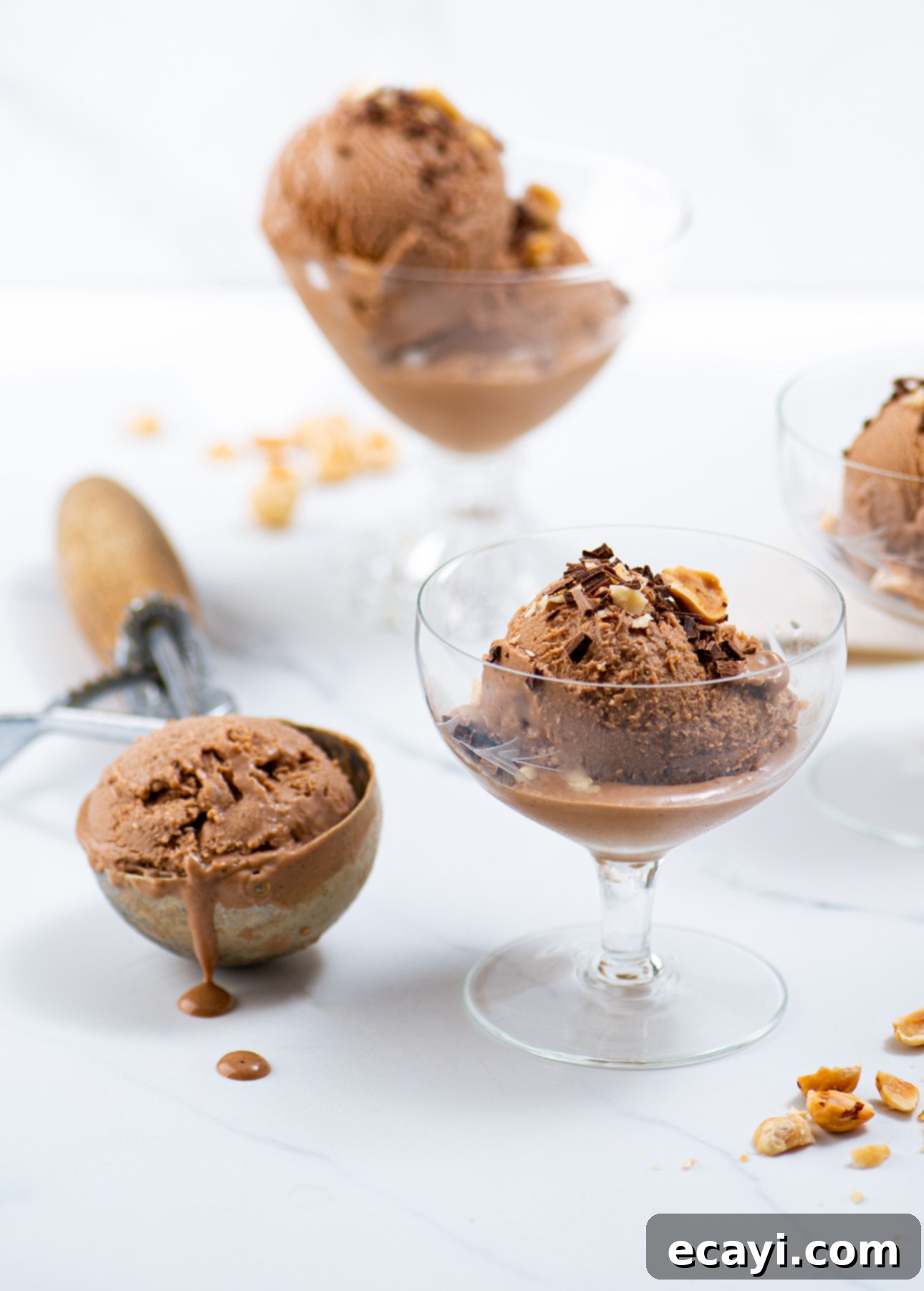 Velvet Chocolate Hazelnut Gelato 2 Scoops of milk chocolate hazelnut gelato in glass cups, garnished with small chocolate shavings and hazelnuts, sit on a white surface. An ice cream scoop with melting gelato is in the background, alongside scattered hazelnuts. // FoodNouveau.com