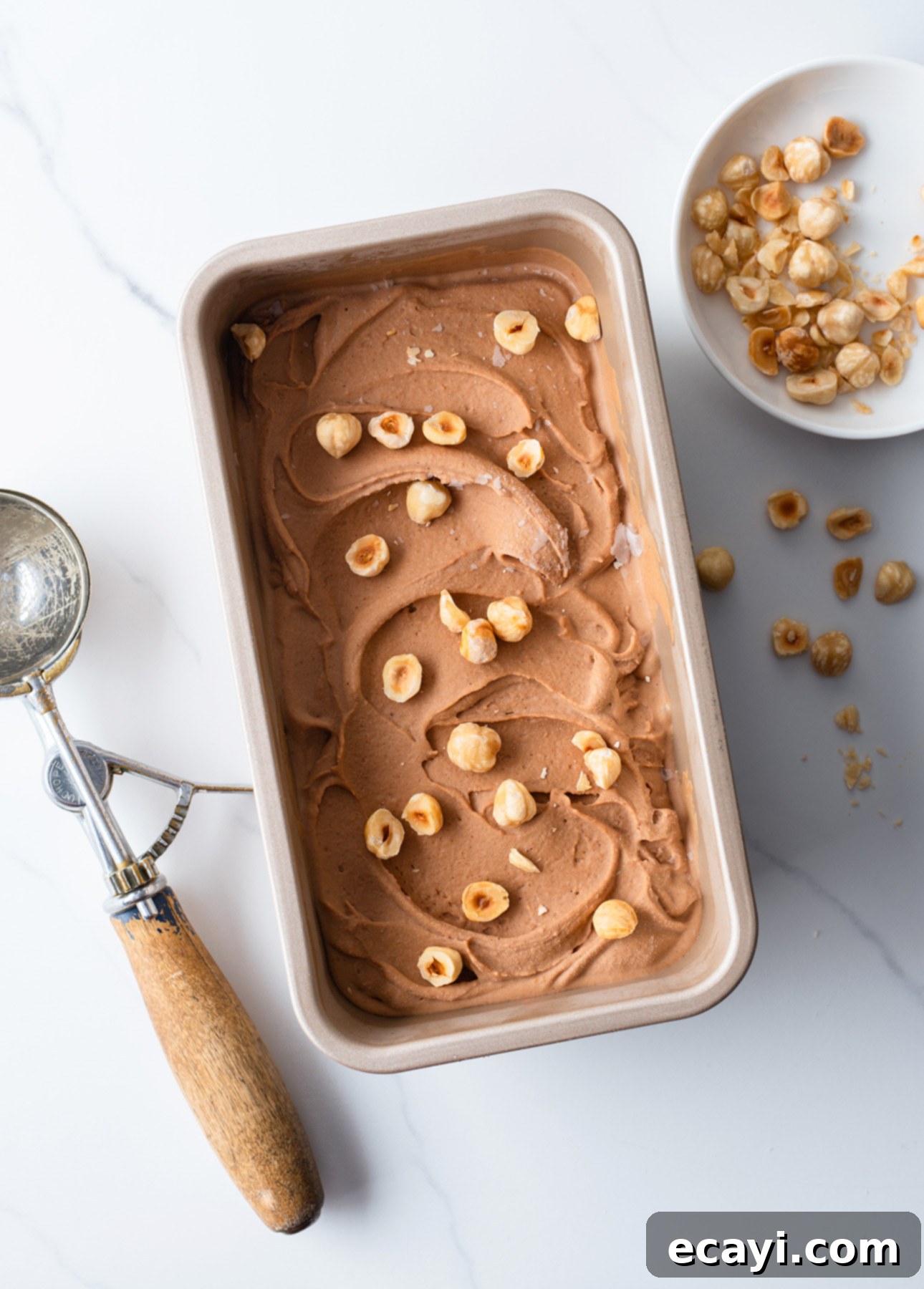 Velvet Chocolate Hazelnut Gelato 3 A rectangular container filled with creamy milk chocolate hazelnut gelato, topped with hazelnuts. A vintage ice cream scoop lies beside it. A small bowl with more hazelnuts is nearby on a white marble surface. // FoodNouveau.com