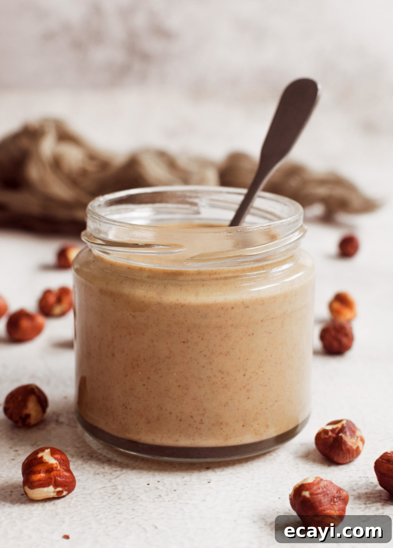 Velvet Chocolate Hazelnut Gelato 4 A jar of smooth, creamy hazelnut butter with a spoon inside. Scattered hazelnuts rest on a textured white surface, while a soft-focus brown cloth adds warmth to the background. // FoodNouveau.com