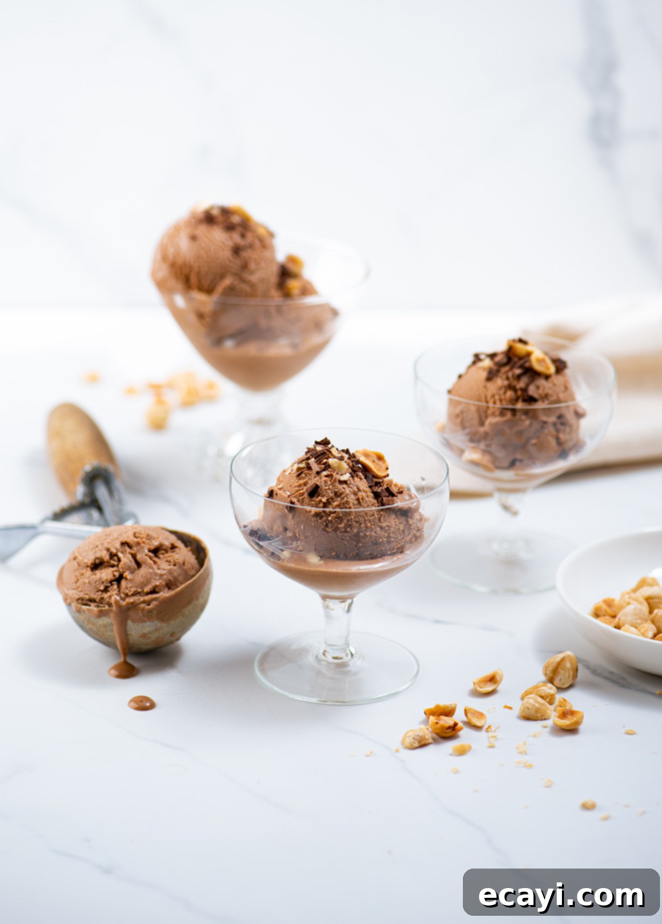 Velvet Chocolate Hazelnut Gelato 6 Three glass cups filled with scoops of milk chocolate hazelnut gelato, crowned with crunchy nuts, rest on a pristine white surface. A metal scoop lies nearby holding a partially melted delight. Scattered nuts and a white bowl complete the tempting tableau. // FoodNouveau.com