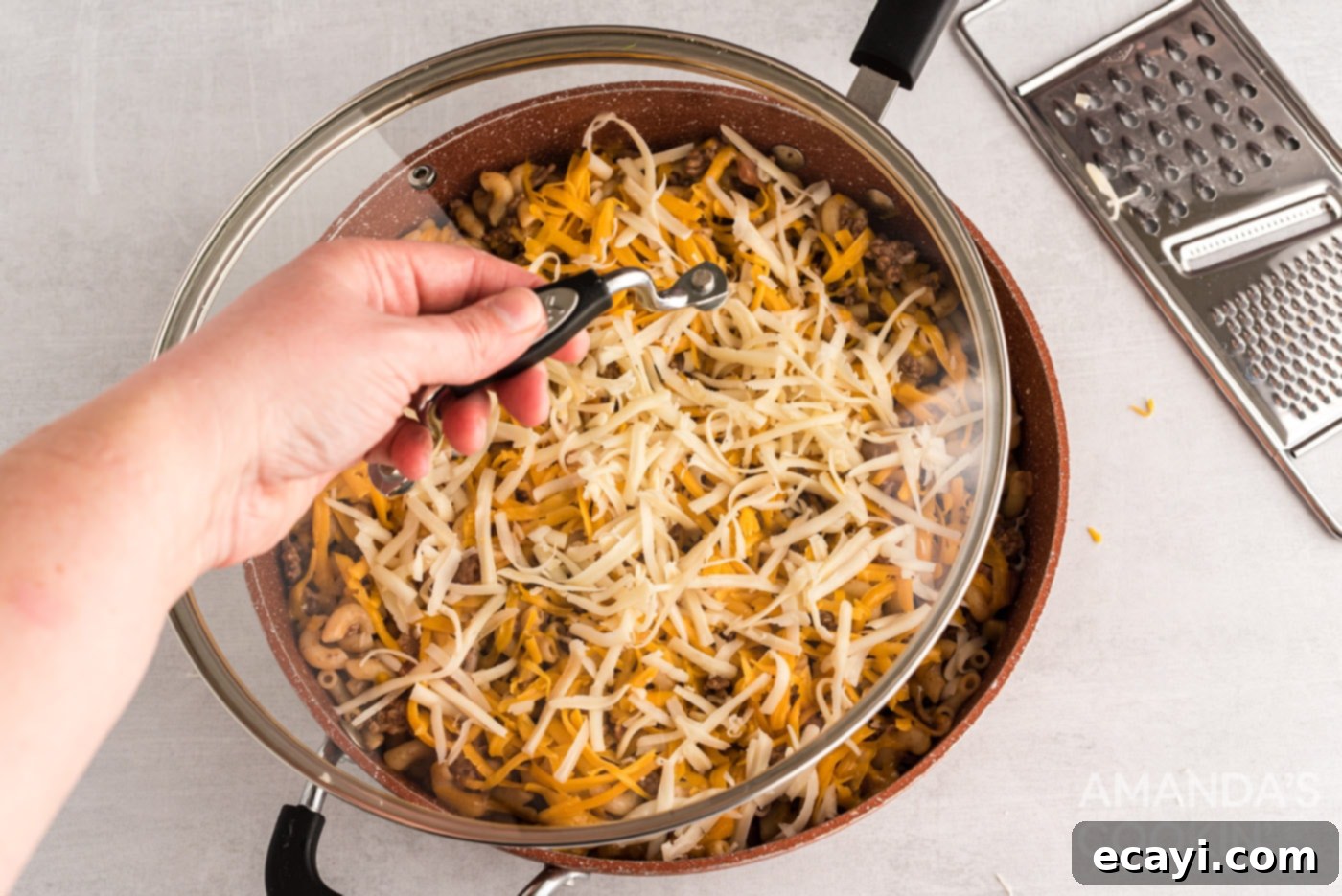 Ultimate Bacon Cheeseburger Bake 9 Shredded cheese being sprinkled evenly over the top of the bacon cheeseburger casserole mixture in a skillet, before melting.
