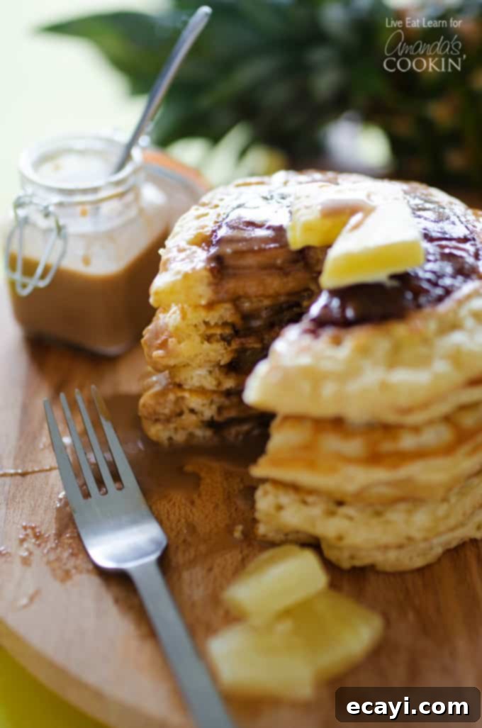 Tropical Pineapple Pancakes with Homemade Coconut Bliss 3 A close up photo of a stack of pineapple pancakes, with one pancake cut to reveal the tender, cooked pineapple inside, on a rustic wooden cutting board.