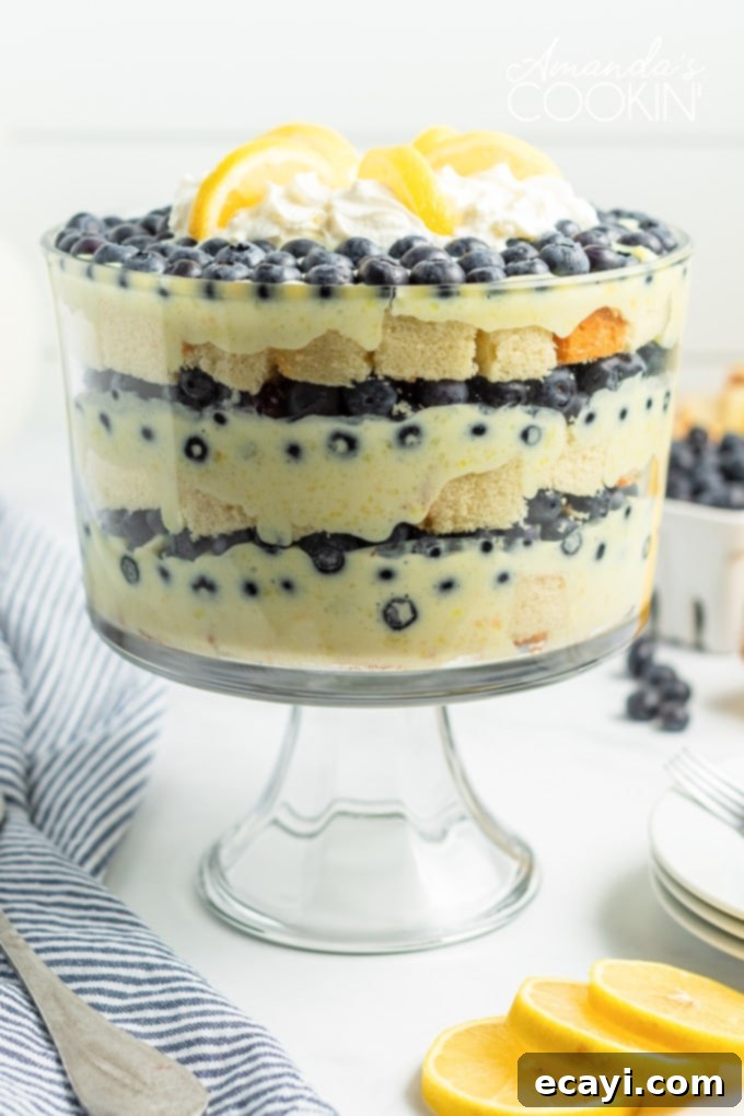 Luscious Lemon Blueberry Trifle: An Easy No-Bake Summer Dessert A beautifully layered lemon blueberry trifle with fresh blueberries and lemon slices on top, resting on a table with a blue towel and whole lemons beside it. The dessert is presented in a clear glass trifle bowl, showcasing its vibrant layers.