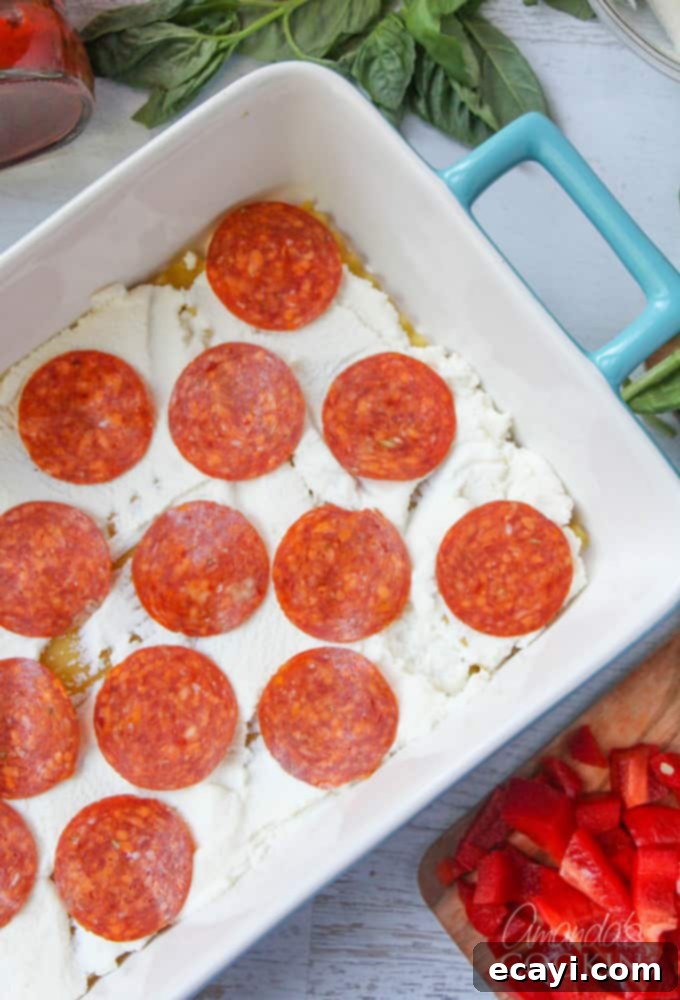 Pizzagna 9 An overhead photo of lasagna noodles in a pan with ricotta cheese spread on top and a vibrant layer of pepperoni slices.