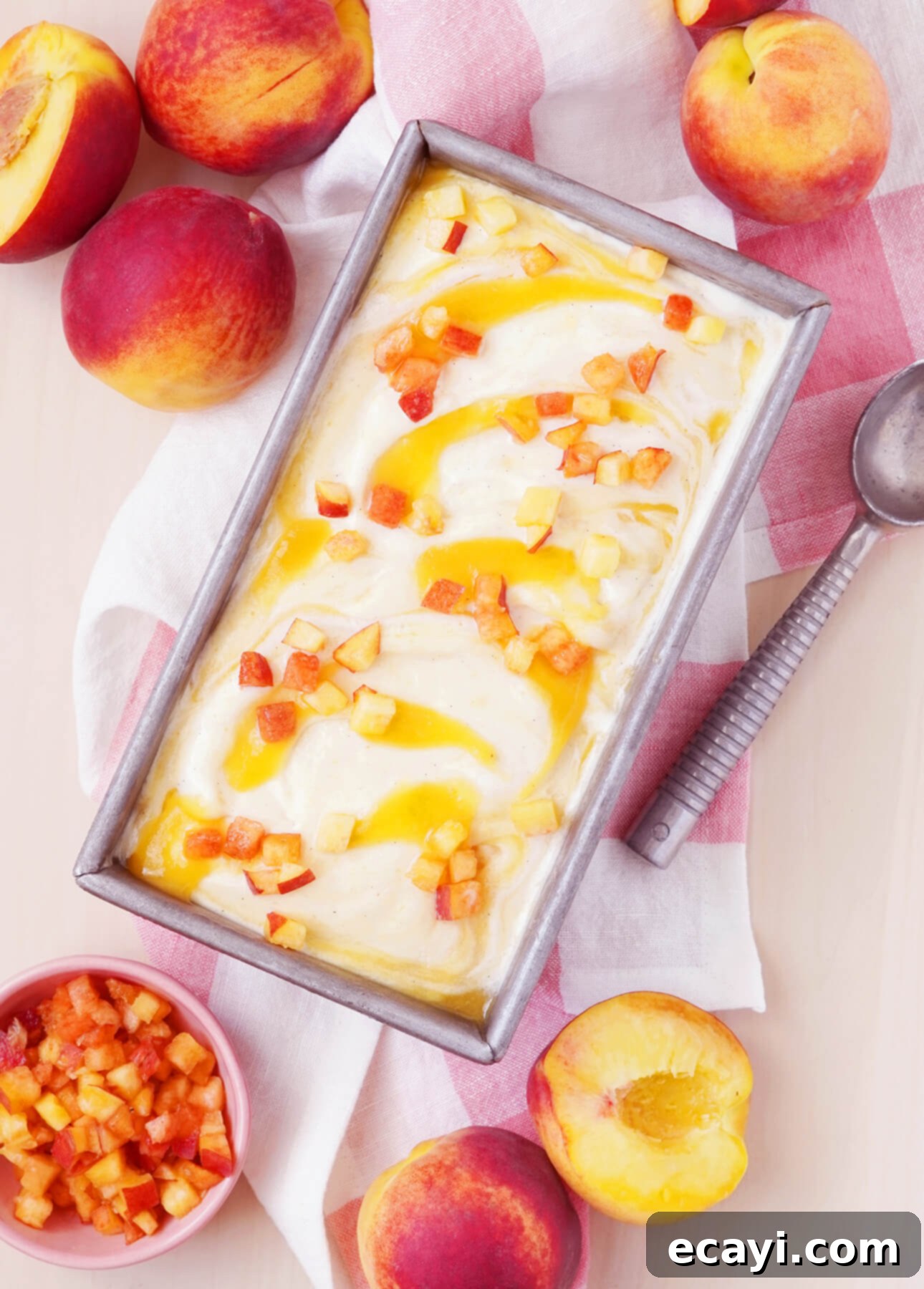 Sun-Kissed Peach Gelato 3 A rectangular metal pan brimming with creamy fresh peach gelato is topped with luscious peach pieces. Fresh whole and halved peaches surround the pan. A pink and white cloth and a metal ice cream scoop are beside it. A bowl of diced peaches is seen in the corner. // FoodNouveau.com