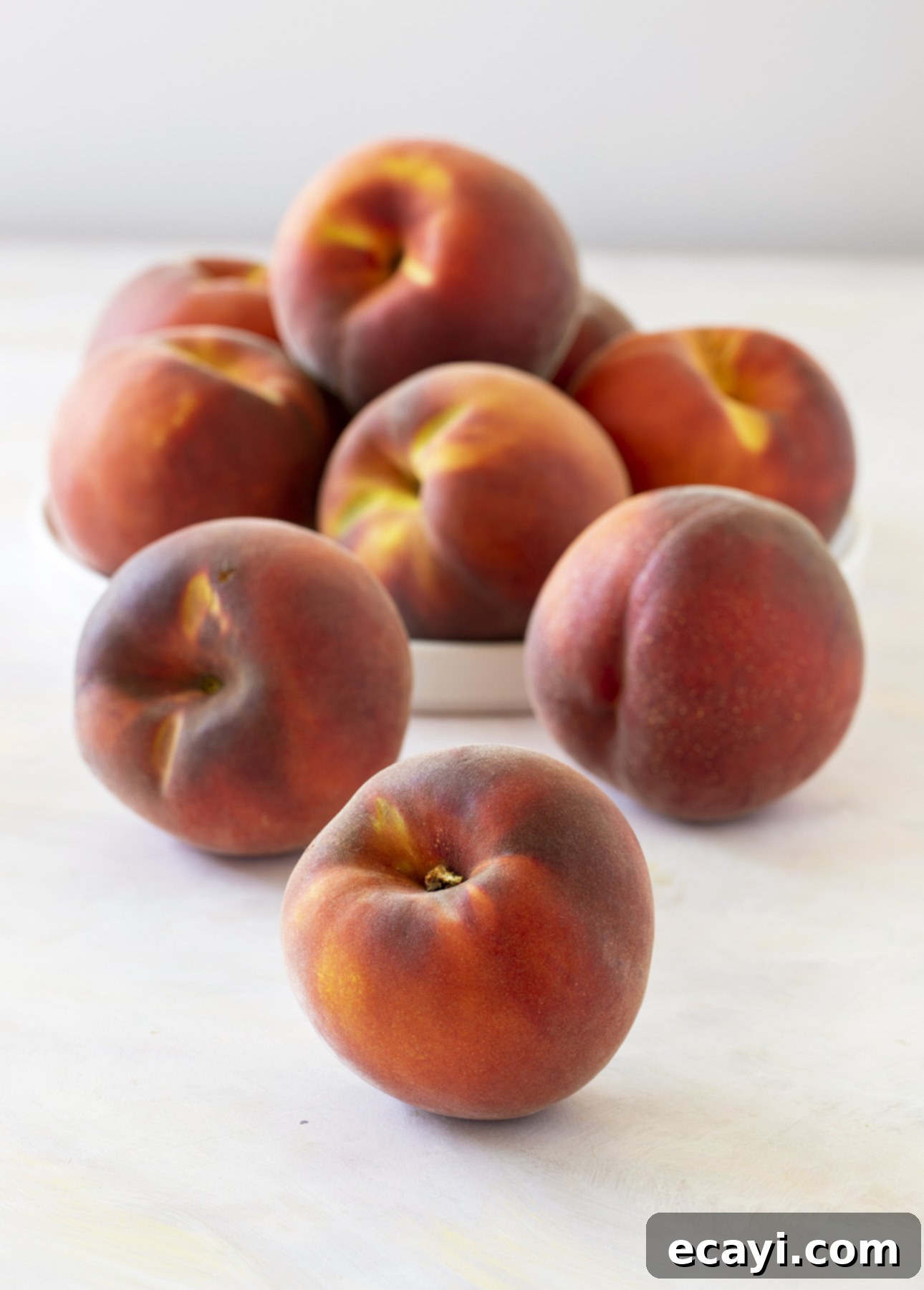 Sun-Kissed Peach Gelato 4 A pile of ripe peaches sits on a light surface. Most peaches are in a white bowl, with a few resting outside. Their reddish-orange hue and velvety texture are accentuated by soft lighting that highlights their freshness. // FoodNouveau.com