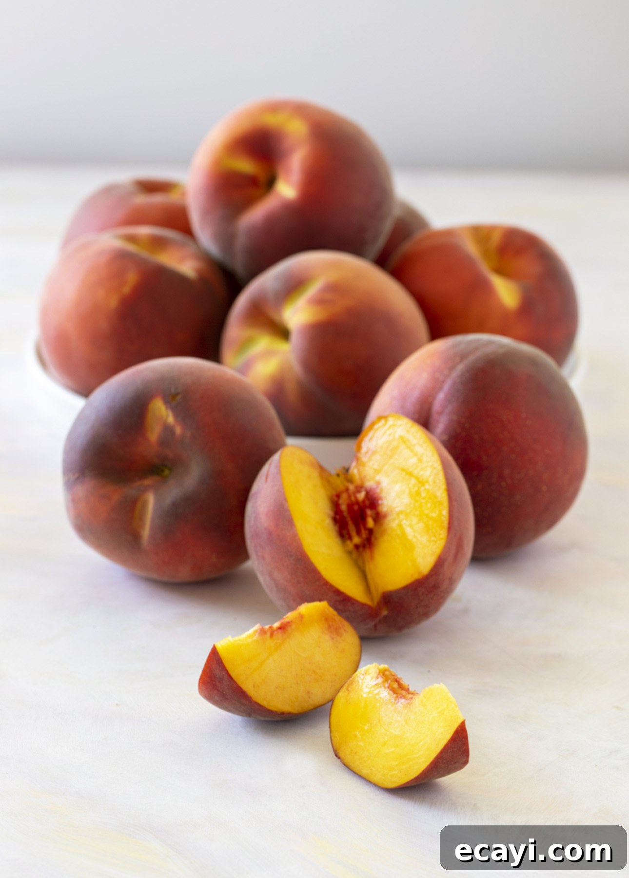 Sun-Kissed Peach Gelato 5 A group of whole peaches is arranged in a bowl, with several more peaches in front. One peach is sliced, revealing its juicy, golden interior against a light background. // FoodNouveau.com