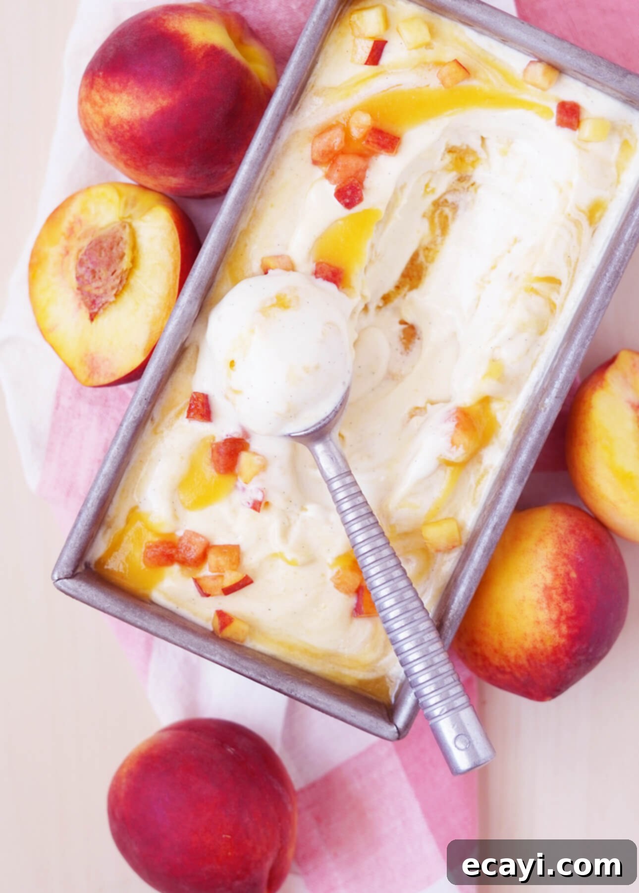 Sun-Kissed Peach Gelato 6 A rectangular tin of fresh peach gelato with a metal scoop nestled inside. The gelato is swirled with chunks of ripe peach, while whole peaches surround the tin on a red and white checkered cloth. // FoodNouveau.com