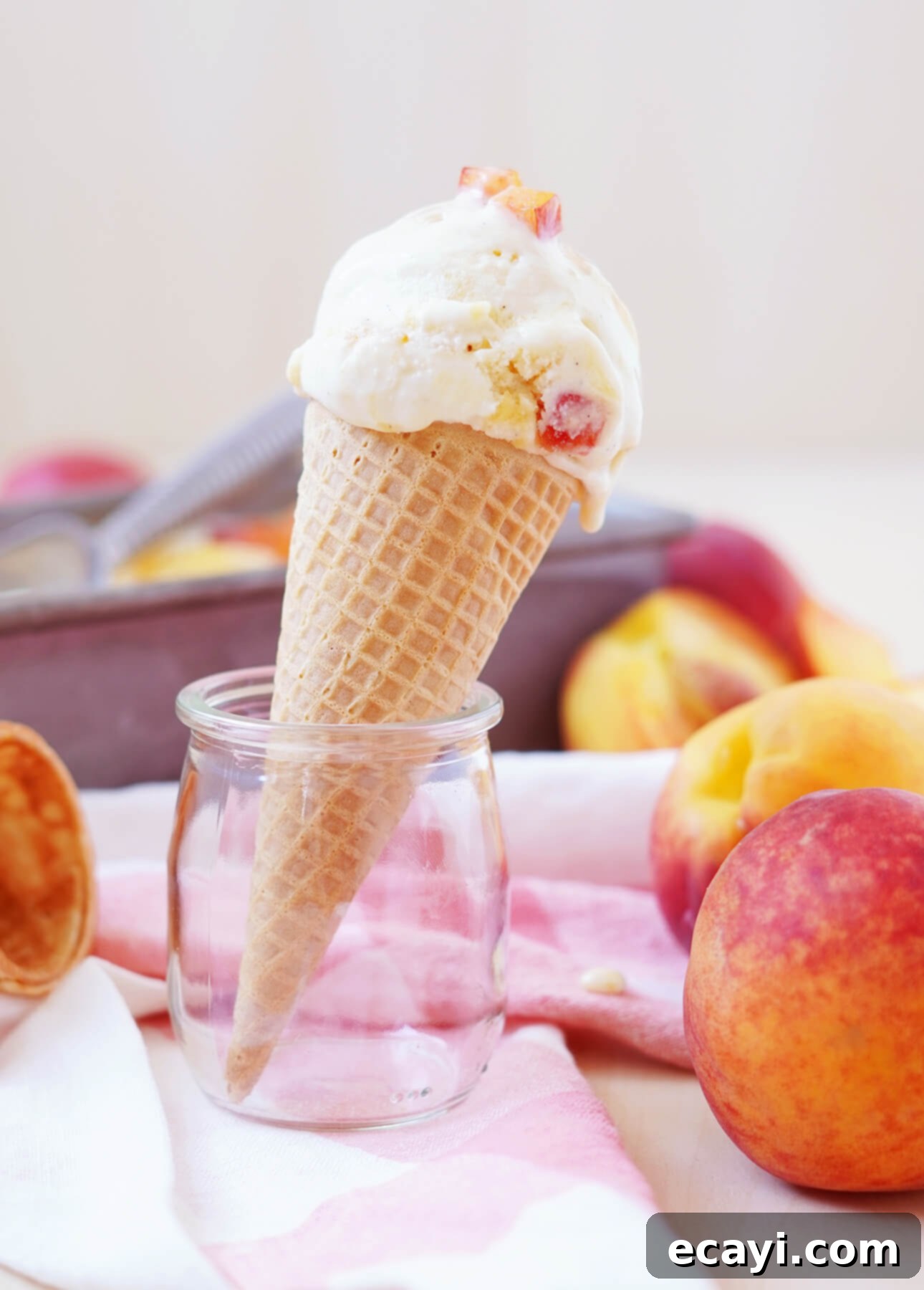 Sun-Kissed Peach Gelato 7 A scoop of fresh peach gelato in a waffle cone balances on a small glass jar. Fresh peaches are scattered around, and a pink-striped cloth lies underneath. A blurred background suggests a sunny kitchen setting. // FoodNouveau.com