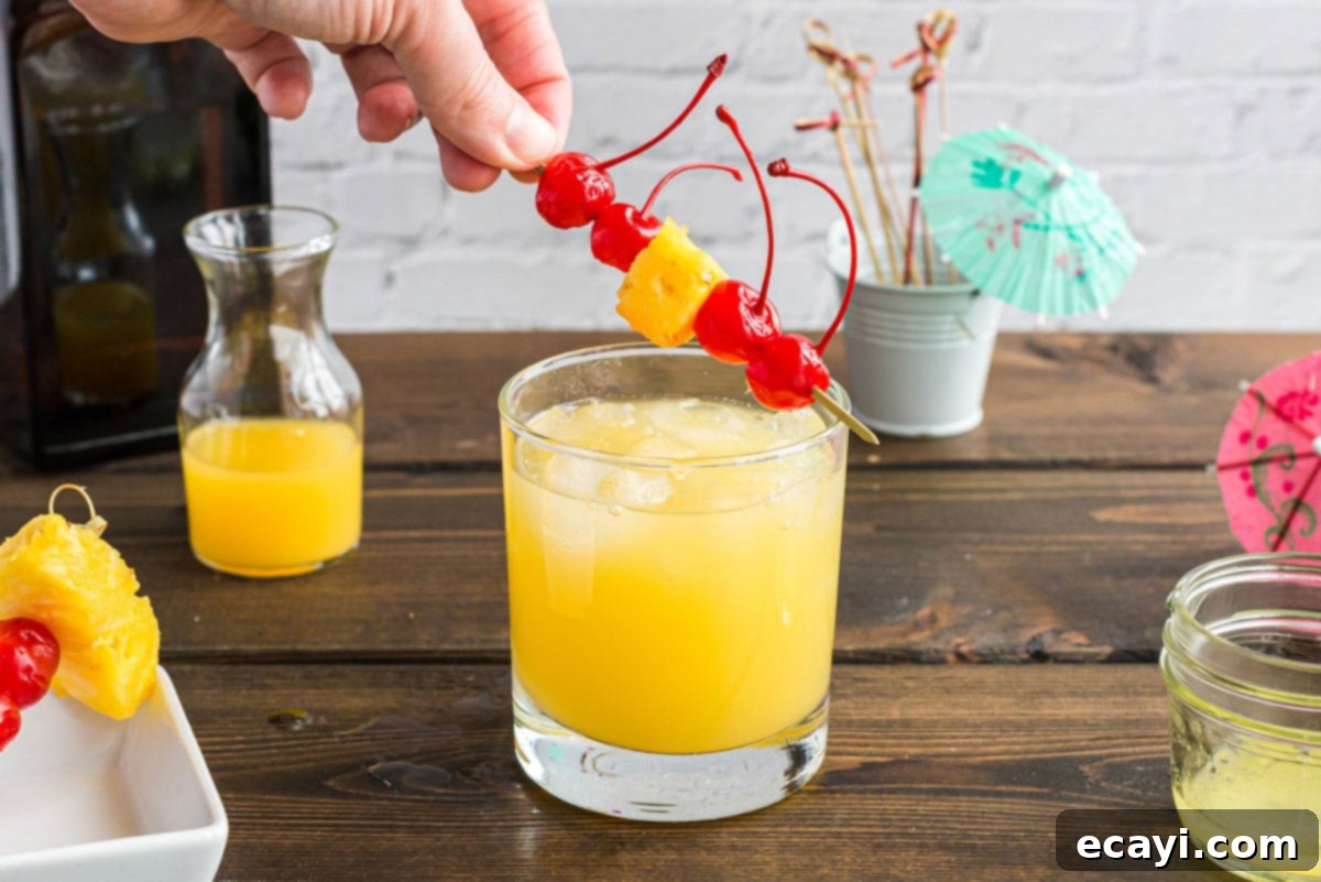 A basic Mai Tai is easy to make. This rum cocktail has combines almond, lime, and tropical fruit flavors. Great for summer and reminds you of the beach! A Mai Tai cocktail in a rock glass being garnished with a vibrant maraschino cherry and a fresh pineapple wedge on a cocktail pick.