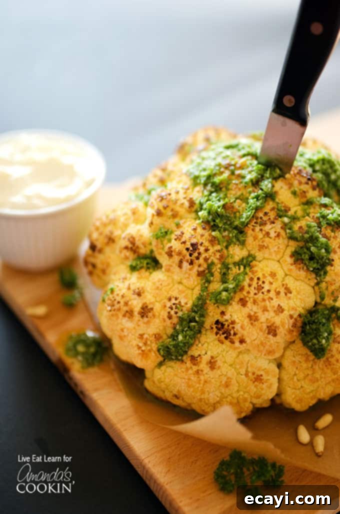 The Ultimate Whole Roasted Cauliflower 3 A close up of a whole roasted cauliflower topped with whipped feta and herb sauce on a wooden cutting board with a knife in the center.