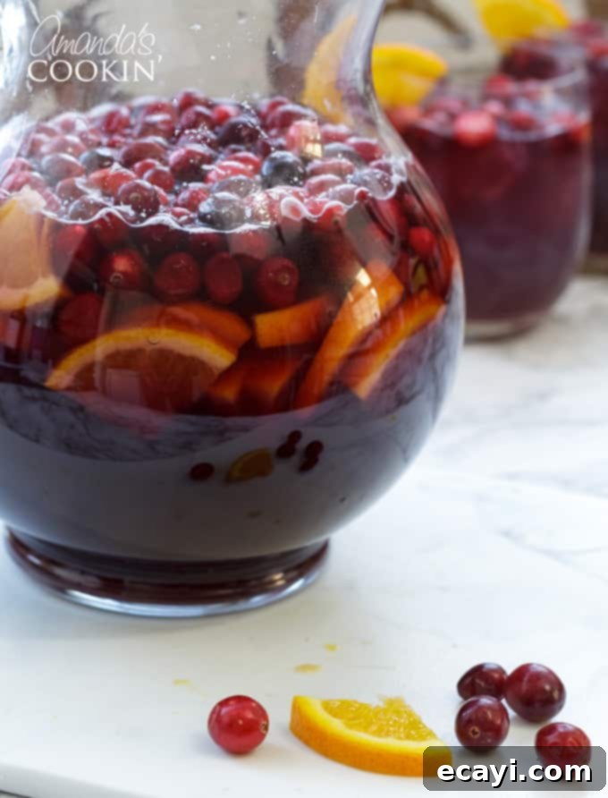 Looking for a great holiday adult beverage? Serve this cranberry sangria to your guests and they will be asking for the recipe and for seconds! #amandascookin #cocktails #sangria #cranberry #orangejuice #christmasrecipes #christmascocktails #thanksgivingcocktails #thanksgivingdrinks A large pitcher filled with cranberry sangria, overflowing with fresh cranberries and orange slices, ready to be poured and enjoyed.