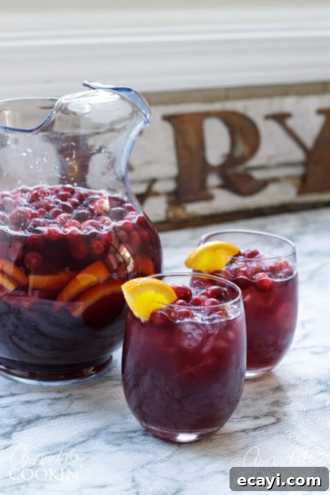 Looking for a great holiday adult beverage? Serve this cranberry sangria to your guests and they will be asking for the recipe and for seconds! #amandascookin #cocktails #sangria #cranberry #orangejuice #christmasrecipes #christmascocktails #thanksgivingcocktails #thanksgivingdrinks A vibrant pitcher of cranberry sangria next to two glasses, garnished with fresh fruit, ready to be enjoyed as a festive beverage.